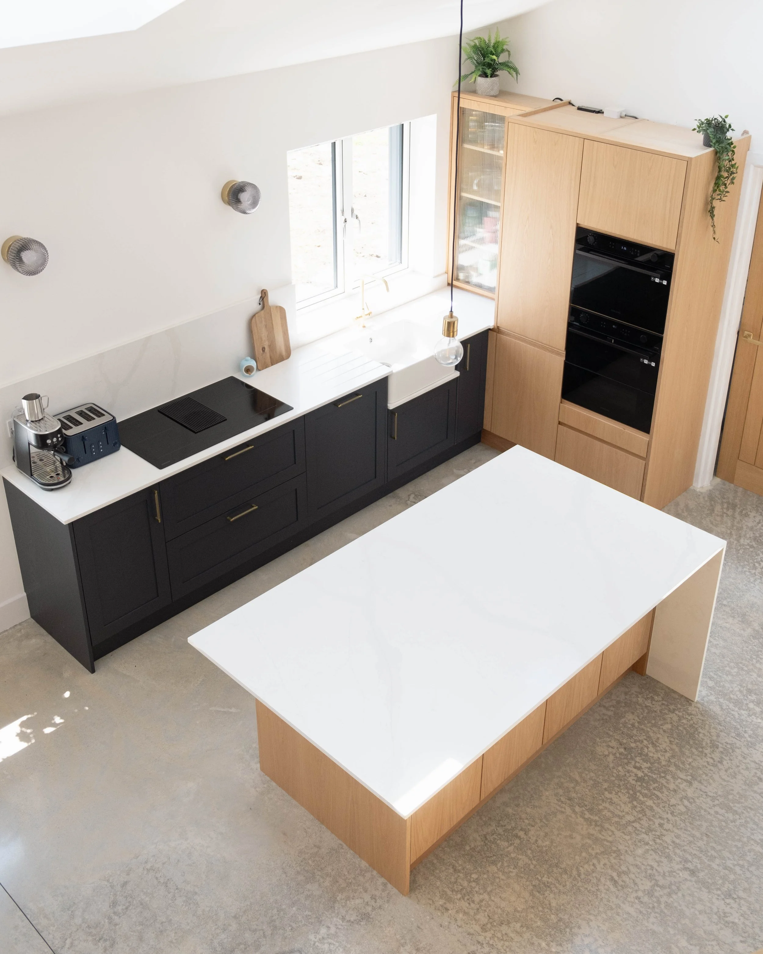 Two kitchen islands with white countertops and wooden bases in a modern kitchen, one with a black stovetop and appliances, and the other with no appliances, in front of a window with natural light. Potted plants and a cutting board are on the wooden 