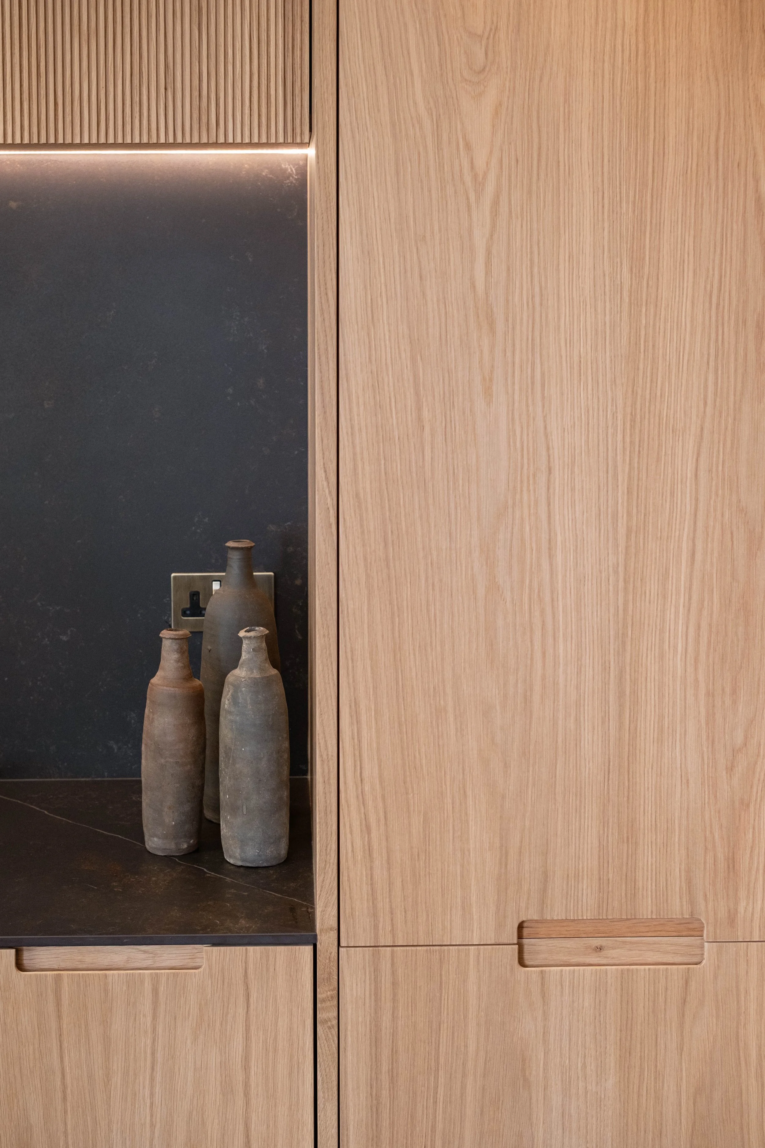 A close-up of wooden cabinets with horizontal handles and a small section of black countertop. Three tall, weathered ceramic vases are positioned on the countertop.