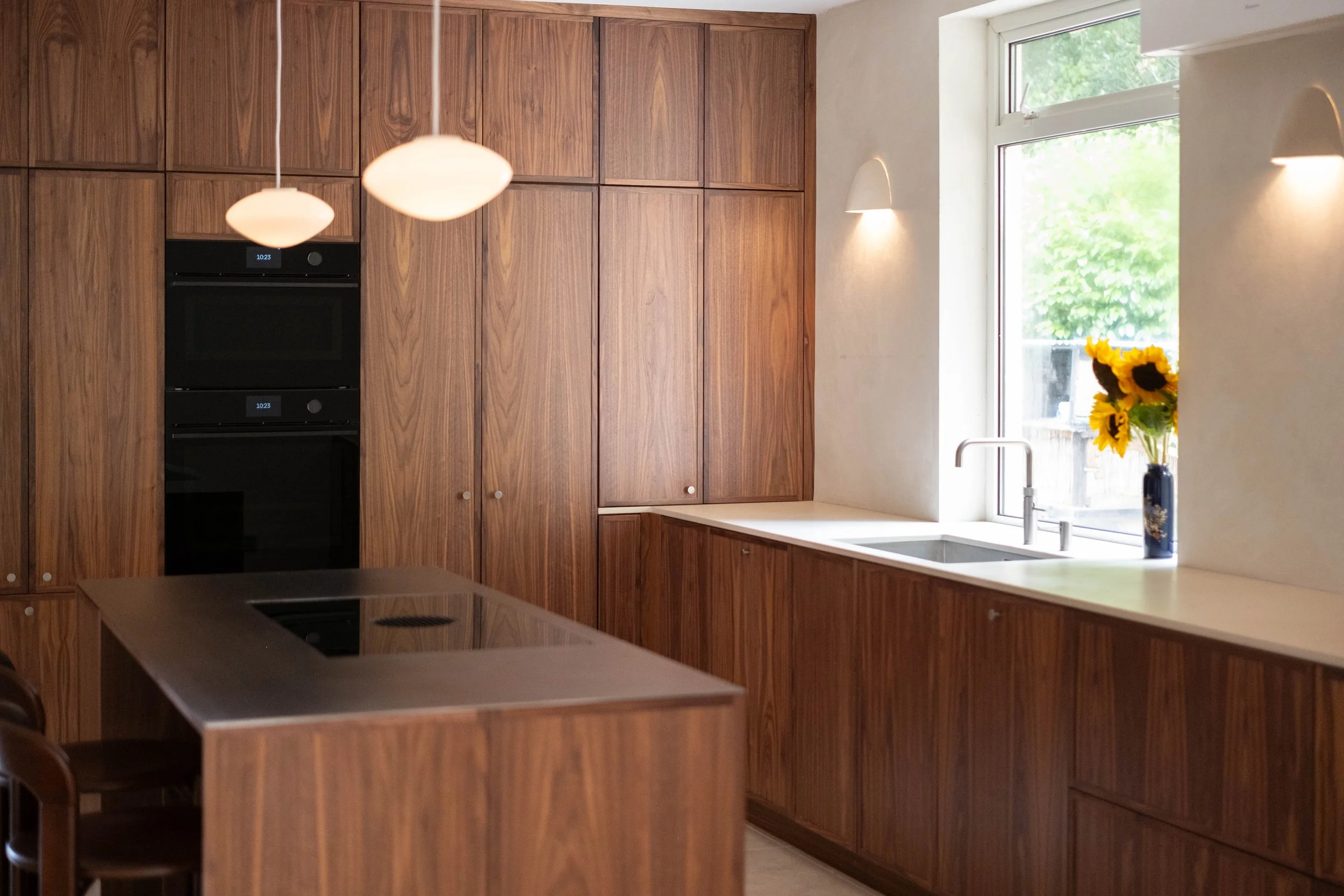 Modern kitchen with wooden cabinets, a window with a view of trees, a vase of sunflowers, and a white countertop with a sink.