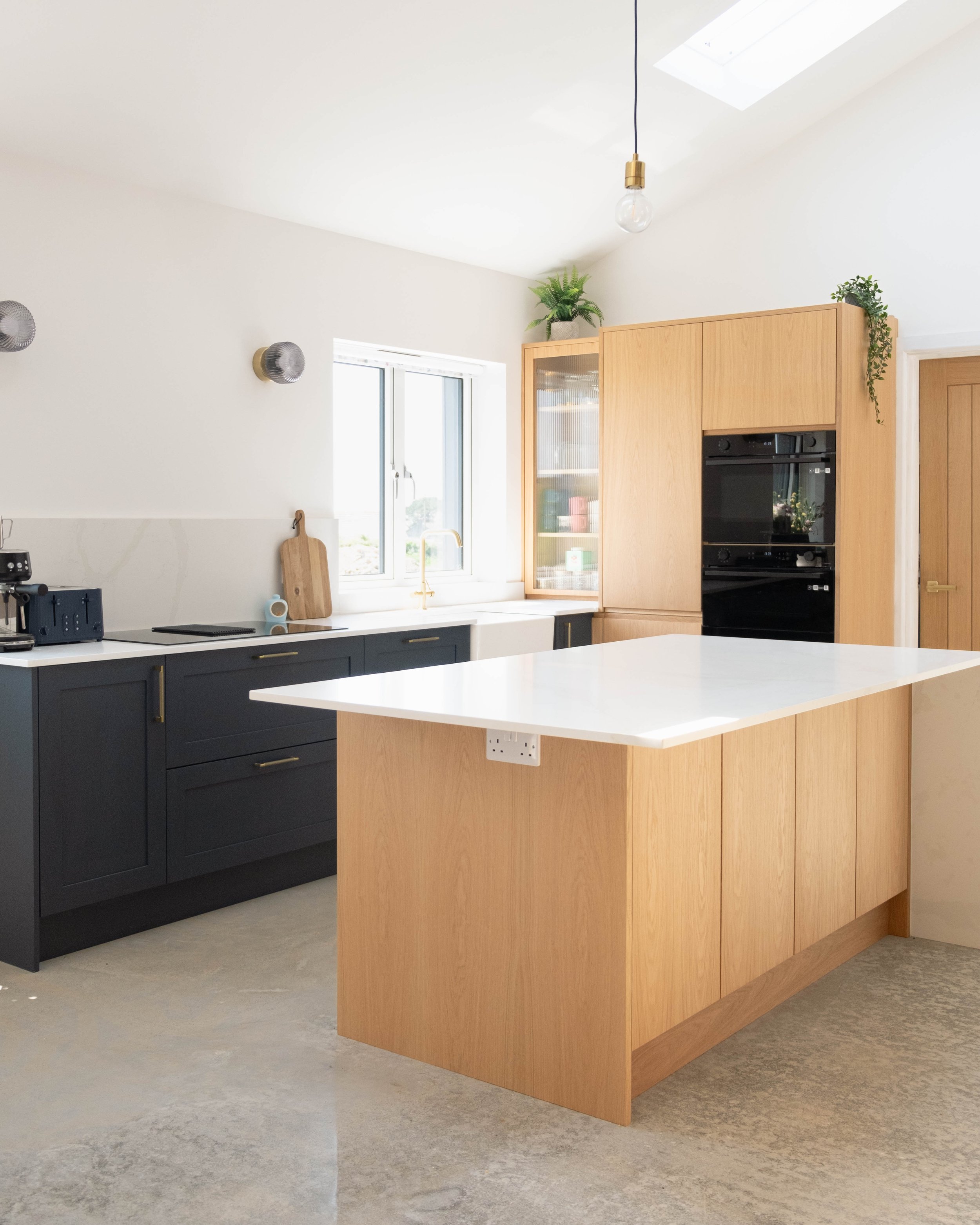 Modern kitchen with natural wood cabinets, black oven, white countertop, and a blue lower cabinet. Decor includes plants on top of cabinets, and a window with a view outside. The ceiling has a skylight and a hanging light bulb.
