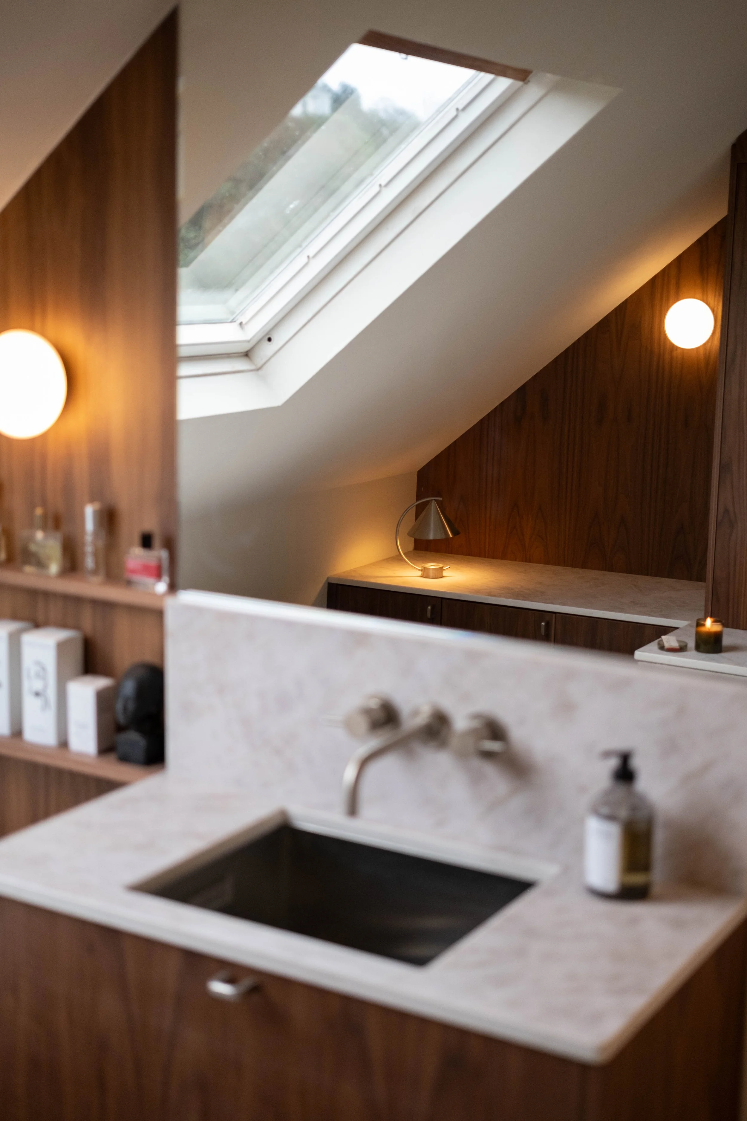 Interior view of a room with a skylight window, wooden wall paneling, and a countertop with a sink and various bottles, illuminated by warm and cool lighting.