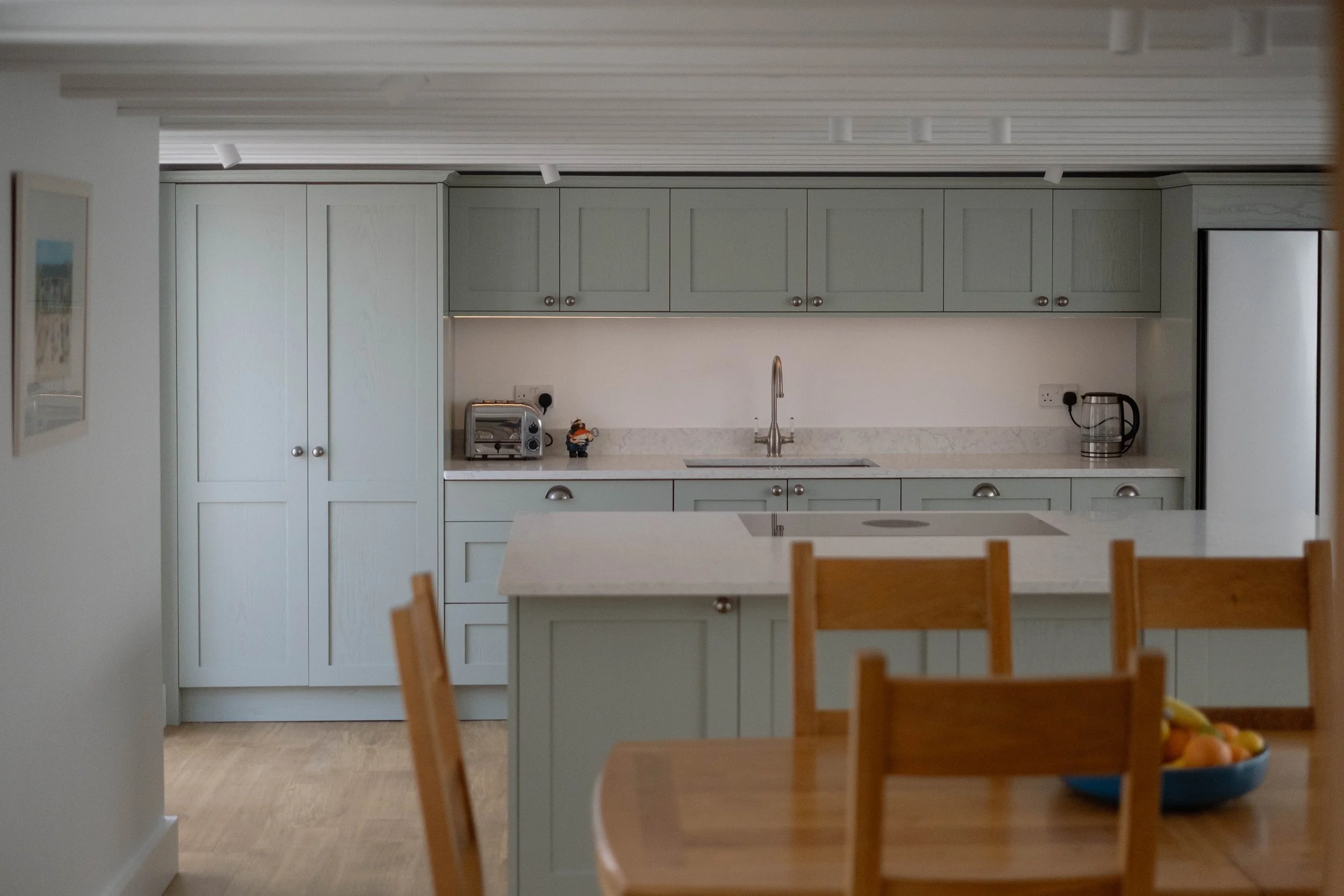 A kitchen with light green cabinets, a marble countertop, and a stainless steel sink. There are small appliances and a decorative figurine on the counter, with a dining table and chairs in the foreground and a fruit bowl on the table.