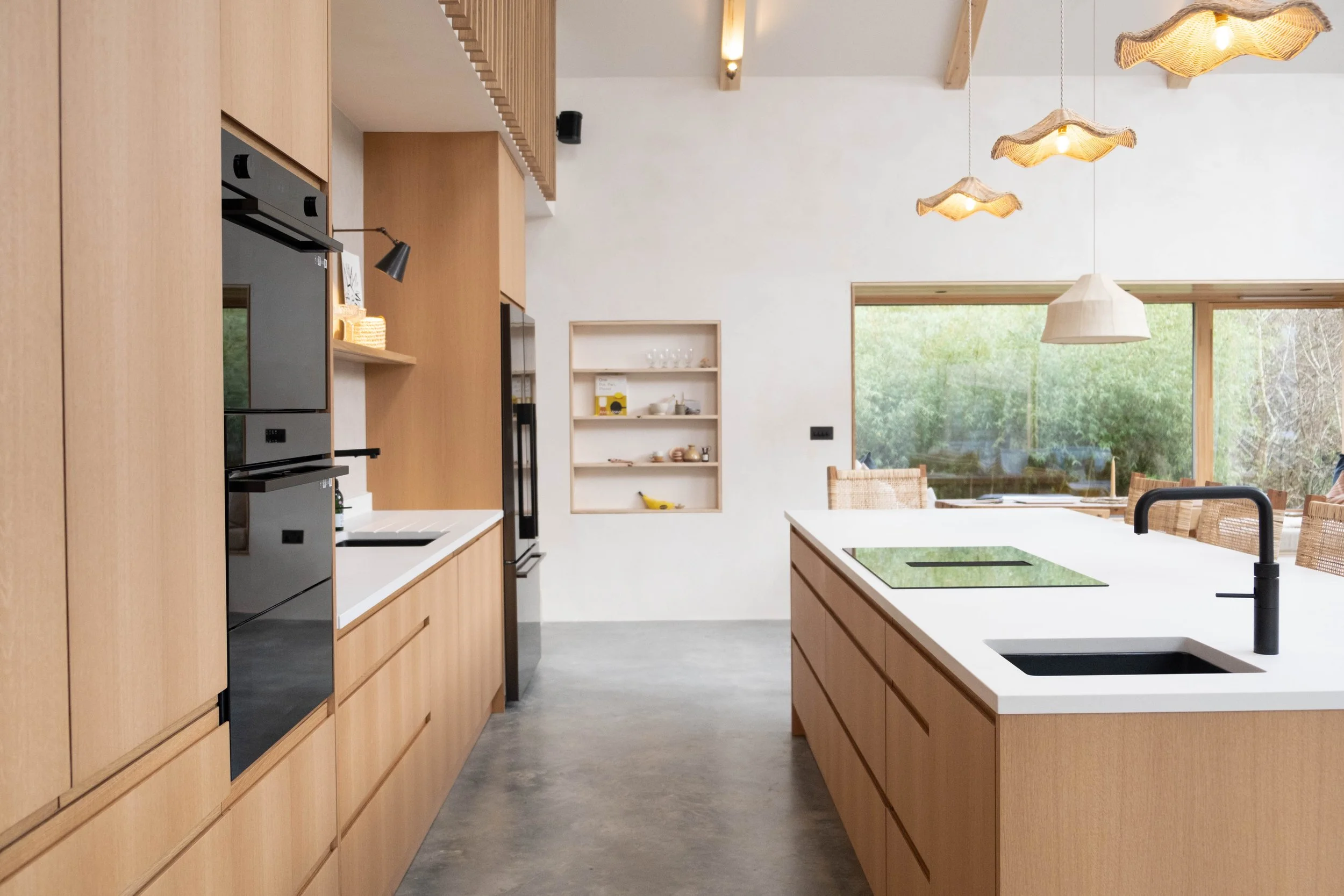 Modern kitchen with wooden cabinetry, white countertops, a large island with a cooktop, black faucet, and large windows overlooking greenery.
