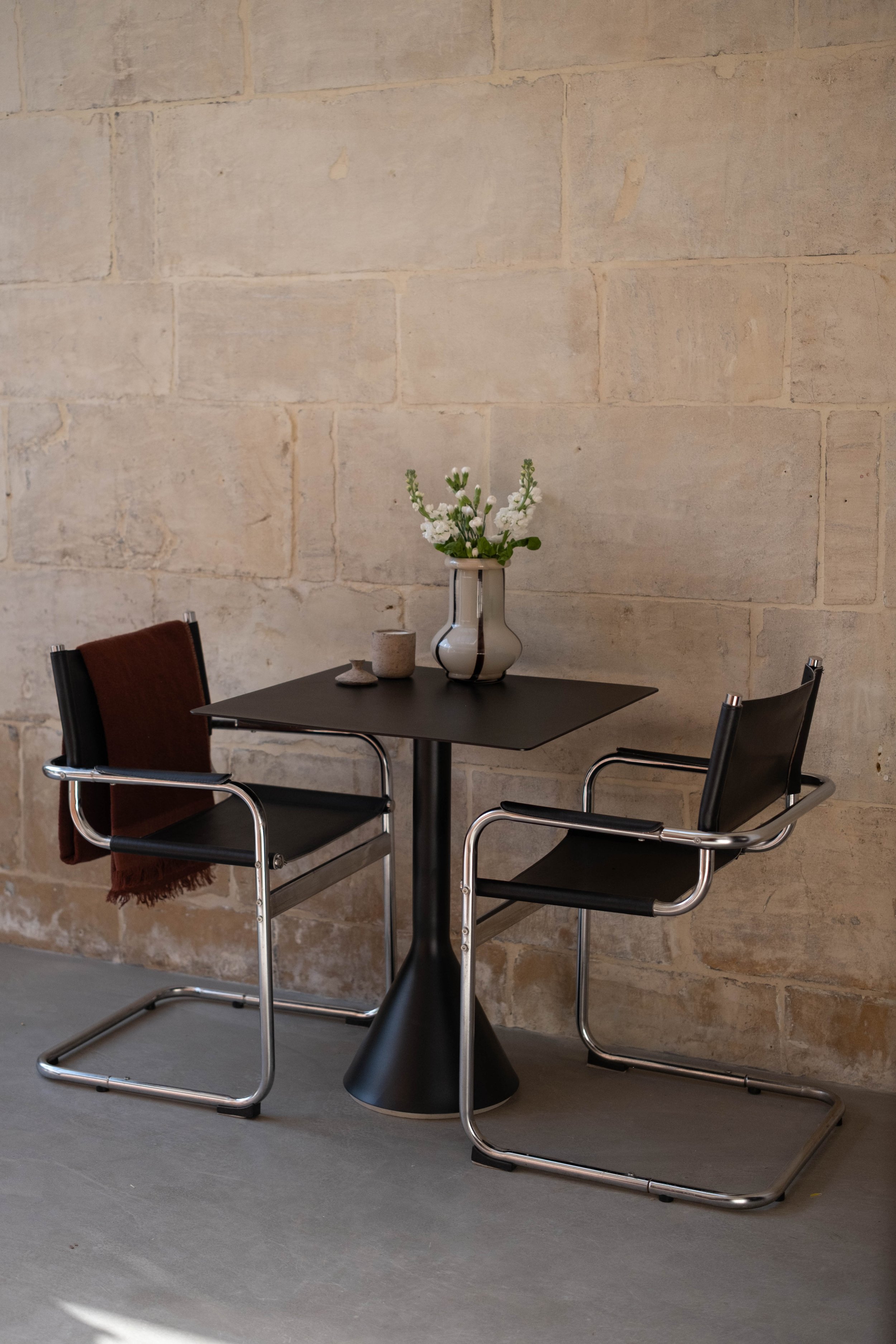 A small black table with a white and black vase of white flowers, two small cups, and two modern black chairs with chrome frames against a beige brick wall.