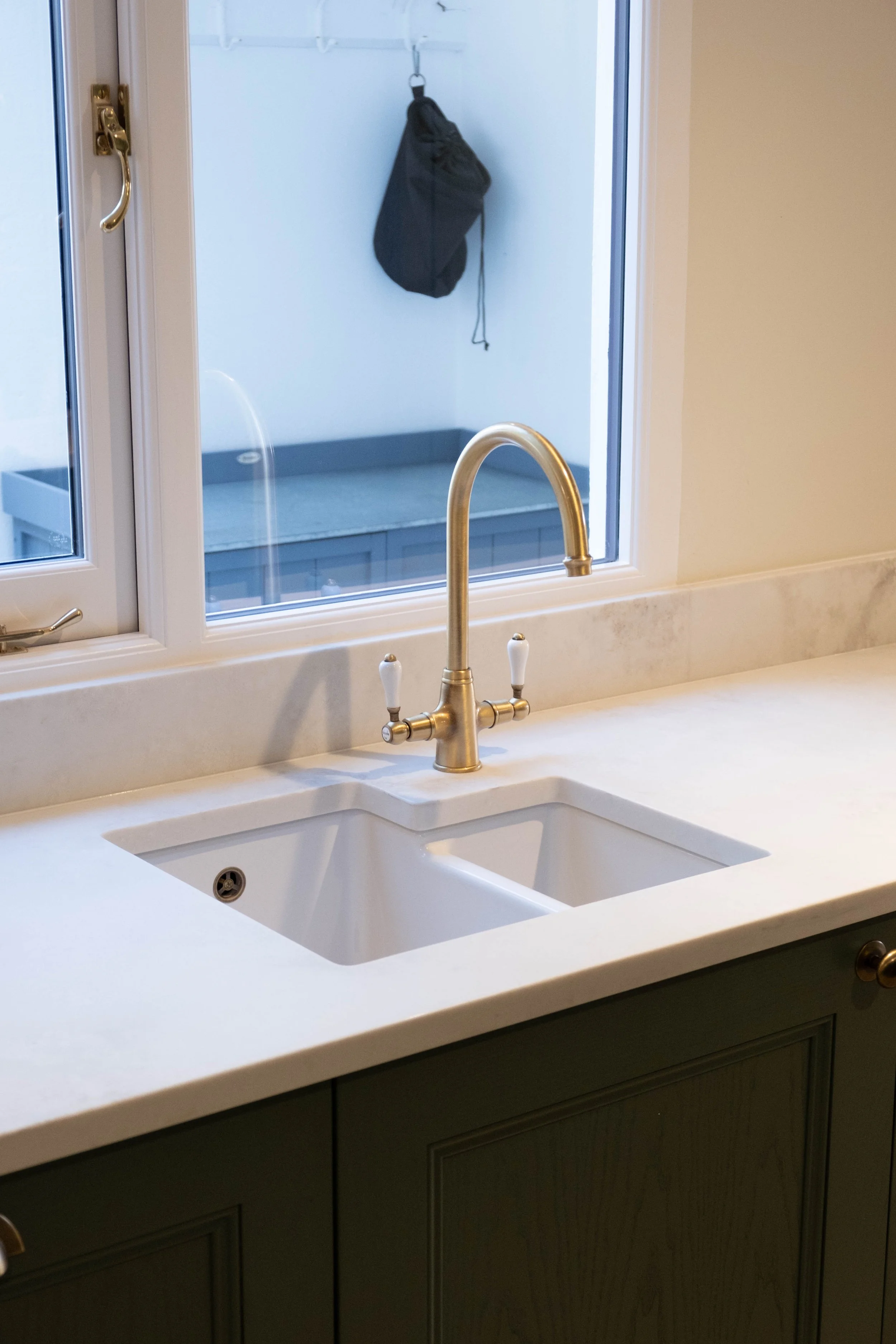 Kitchen sink with brass faucet, white countertop, window above, view of closet with black bag hanging inside.