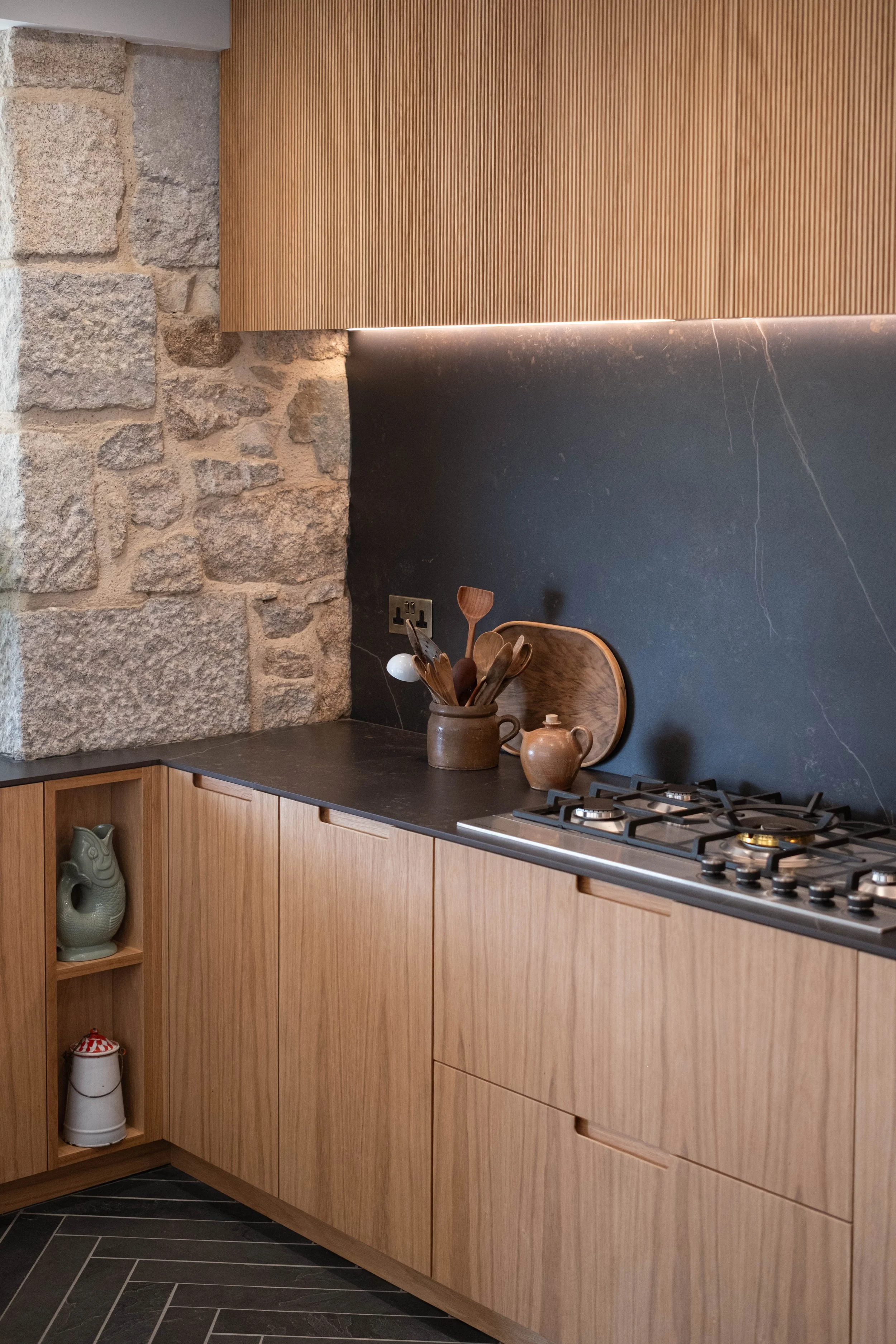 A kitchen corner with a wooden cabinet, black countertop, stone wall, and decorative items including pottery and wooden utensils.