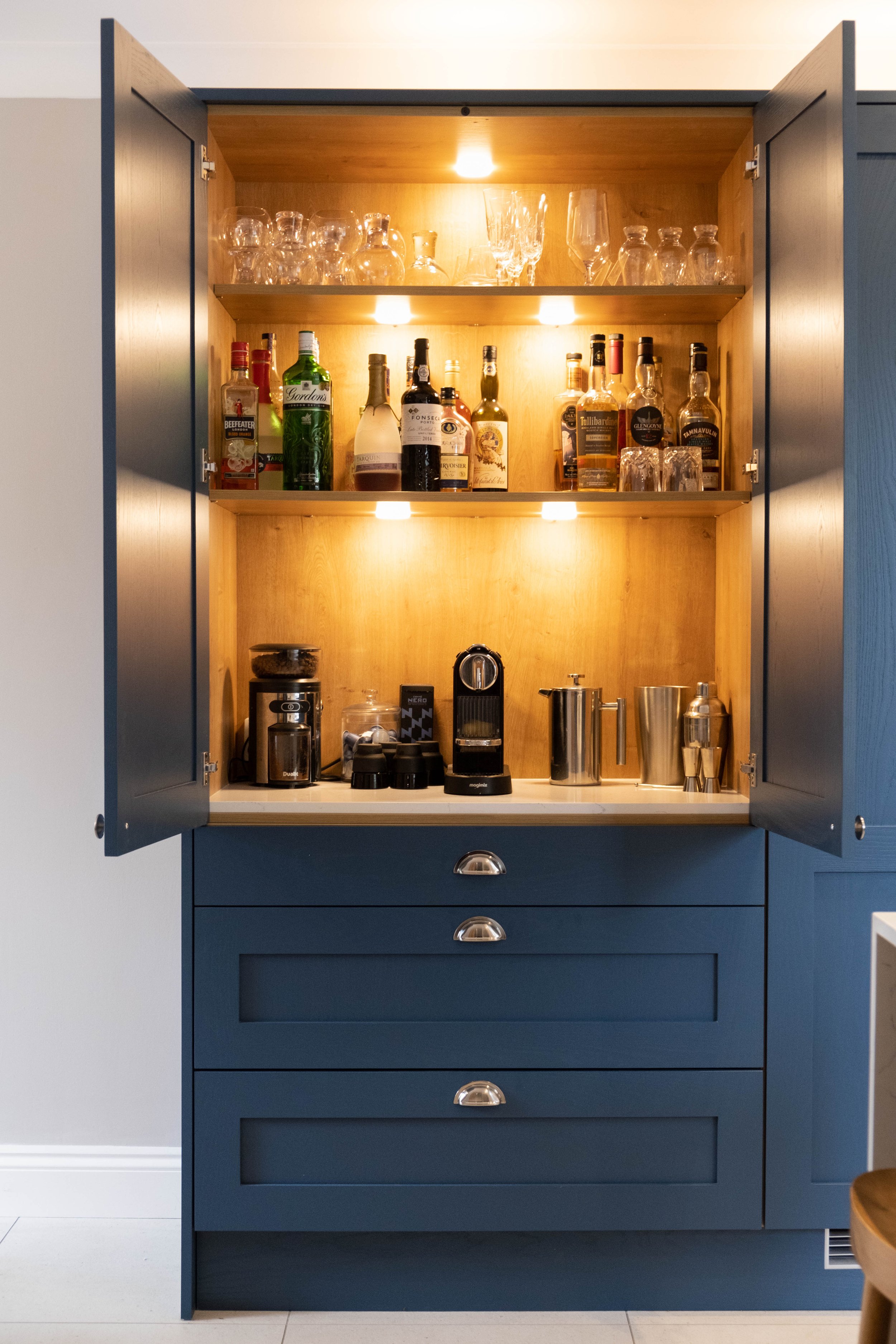A blue kitchen cabinet with glassware and liquor bottles on open shelves, under warm lighting, with a white counter and drawers below.