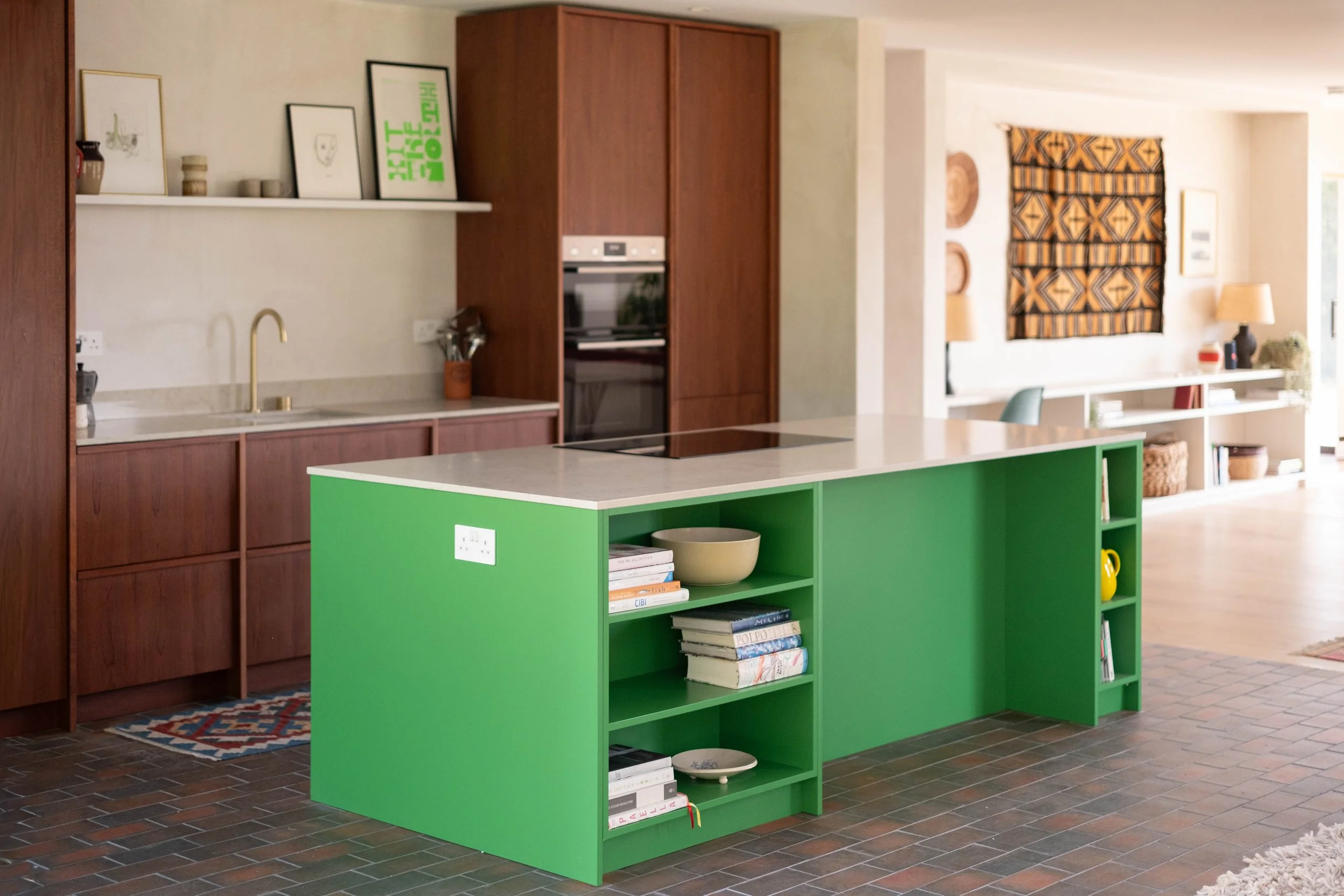 A kitchen with a green island, wooden cabinets, a wall shelf with framed pictures, and a living room area with a wall hanging, lamps, and shelves with books and decor.