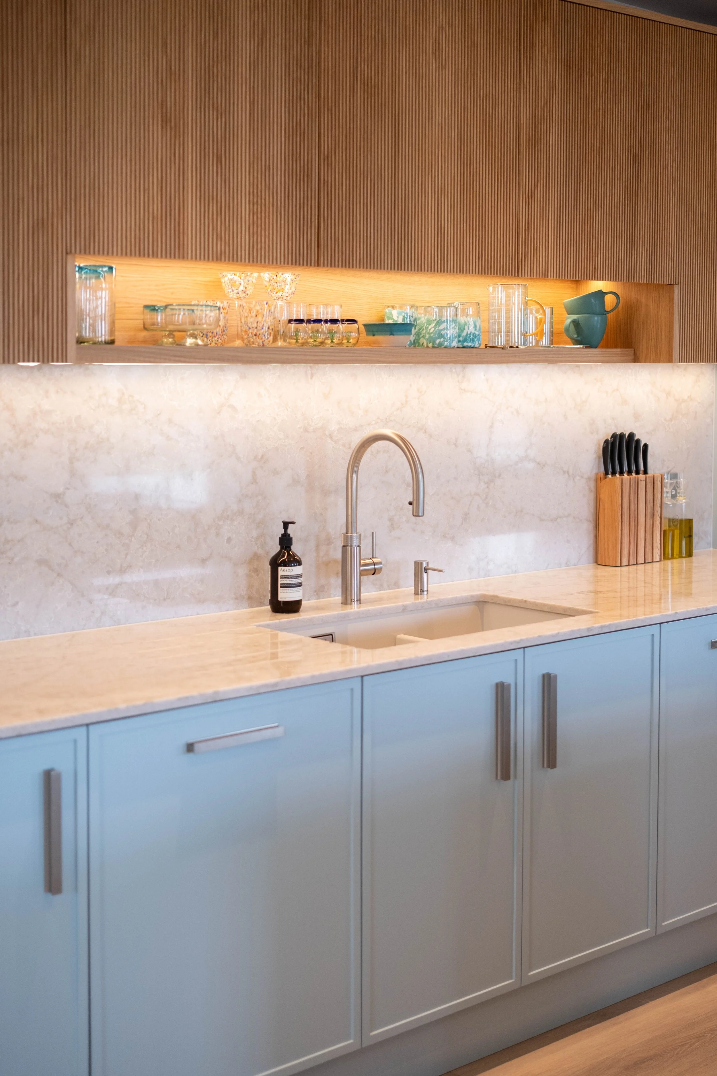 Kitchen with light blue cabinets, white marble countertop, and a beige backsplash. Open wooden shelf above the counter holds glassware and cups. Sink with chrome faucet in the center, soap dispenser on the counter, and a knife block with knives on th