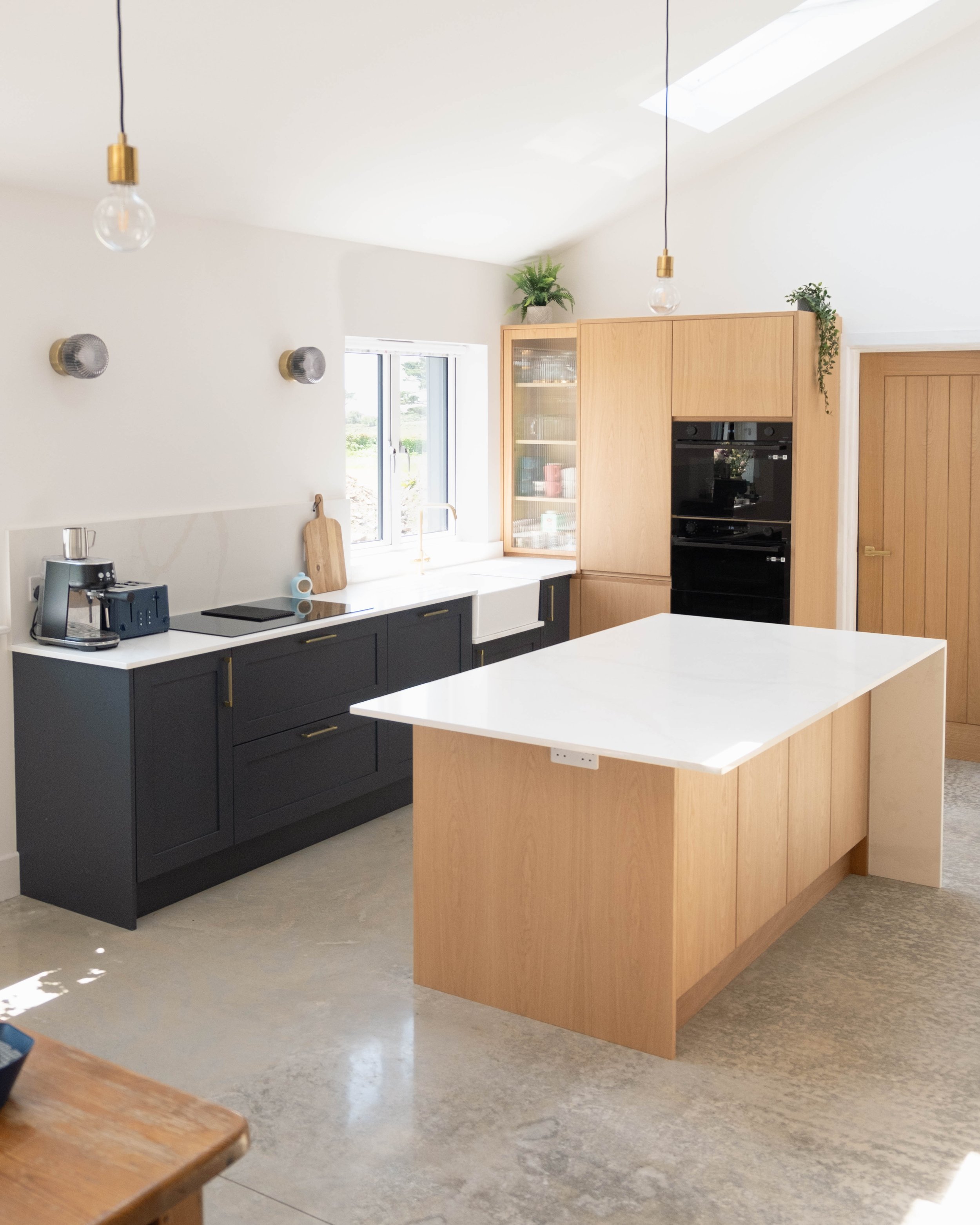 Modern kitchen with black and wood cabinetry, white countertops, and stainless steel appliances. Pendant lights hang from the ceiling, and a window provides natural light.