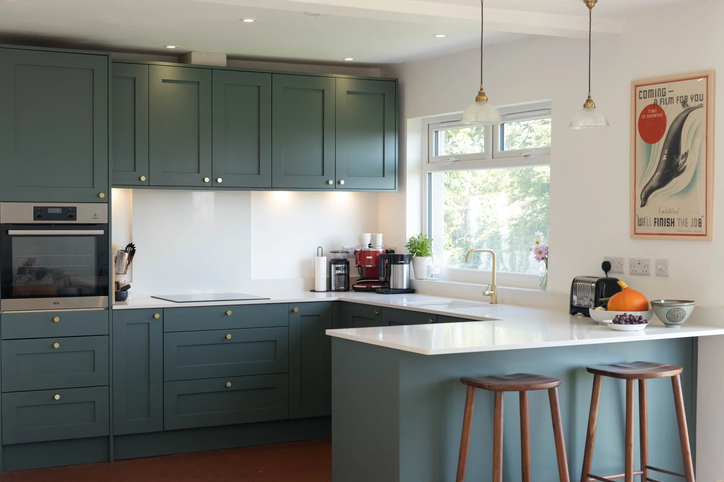 Modern kitchen with teal cabinets, white countertops, and a window with green trees outside, decorated with a vintage movie poster on the wall and various small kitchen appliances on the counter.