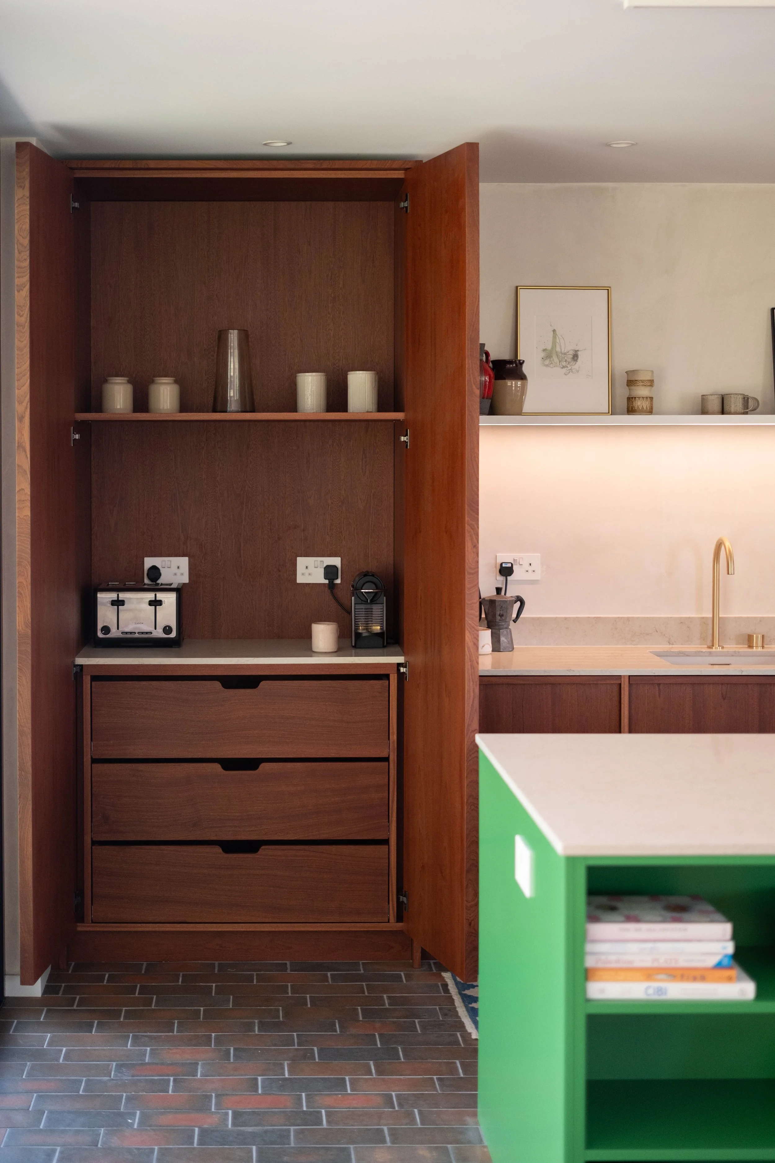 A wooden cabinet with three drawers at the bottom and open shelves at the top, with various vases and jars. Next to it is a kitchen counter with a built-in sink and a faucet, a small appliance, and a framed picture on the wall. The floor is tiled with rectangular brown tiles.