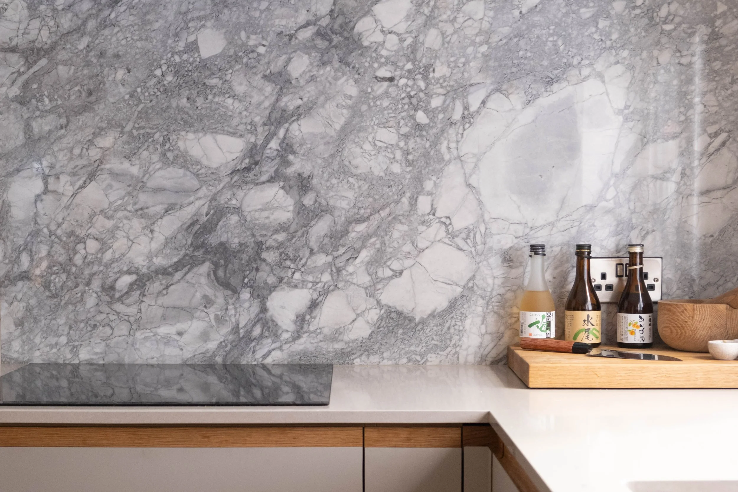 A kitchen countertop with a gray marble backsplash and some bottles, a wooden cutting board, and a small bowl.