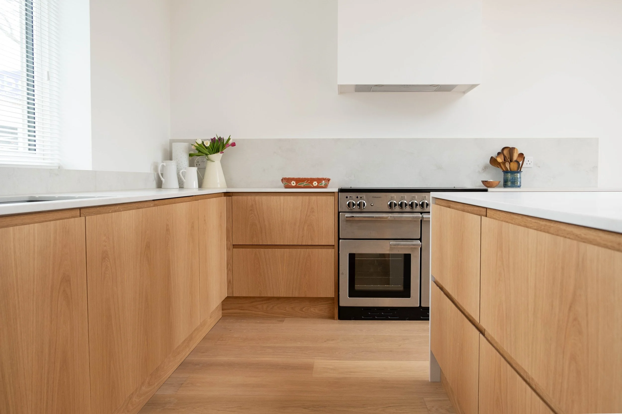 Modern kitchen with wooden cabinets, stainless steel oven, white countertops, a vase with flowers, and utensils in a jar.