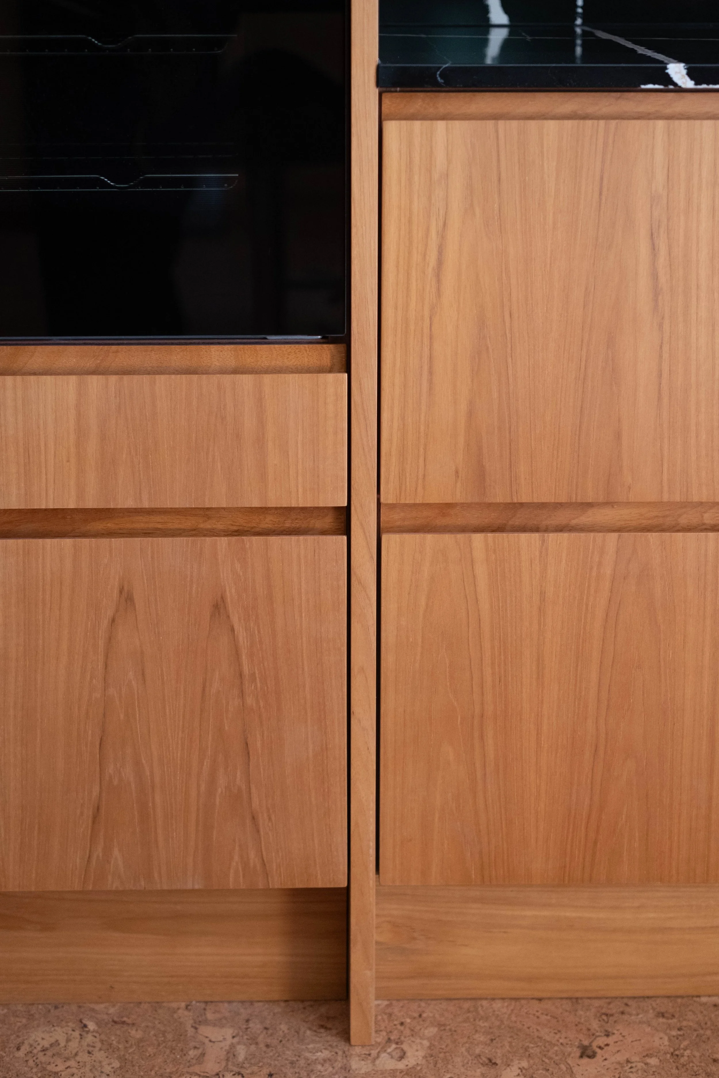 A close-up view of wooden cabinetry with a black stove or oven on the left and light wood cabinets with a vertical split in the middle. The floor is textured and light-colored.