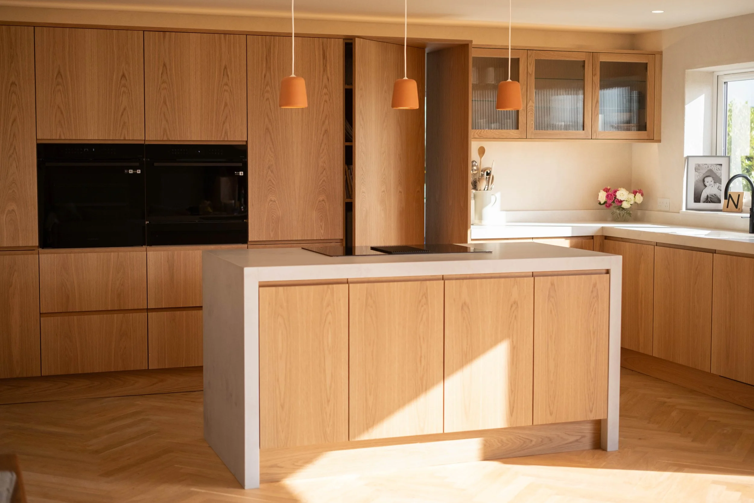 Modern kitchen with wood cabinetry, white countertop island, pendant lights, and a window with a framed black and white photo and flowers