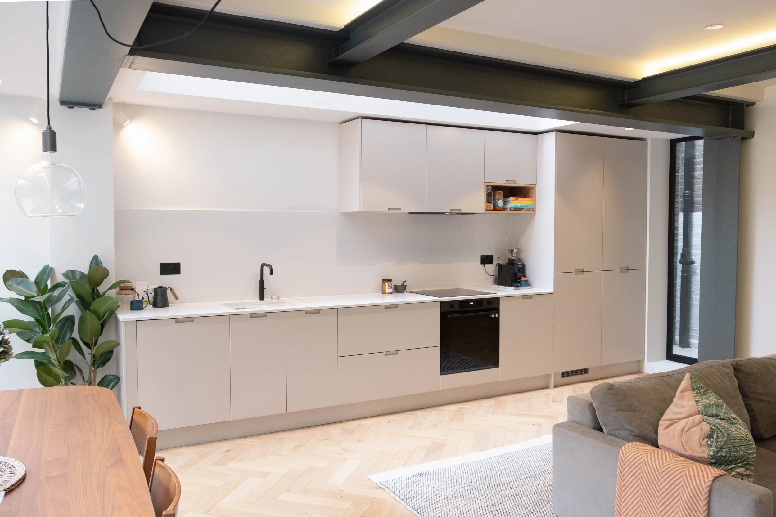 Modern kitchen with white cabinetry, black appliances, black faucet, and open shelving containing jars and books, adjacent to a dining area with wooden table and chairs, a green plant, and a gray sofa with cushions.