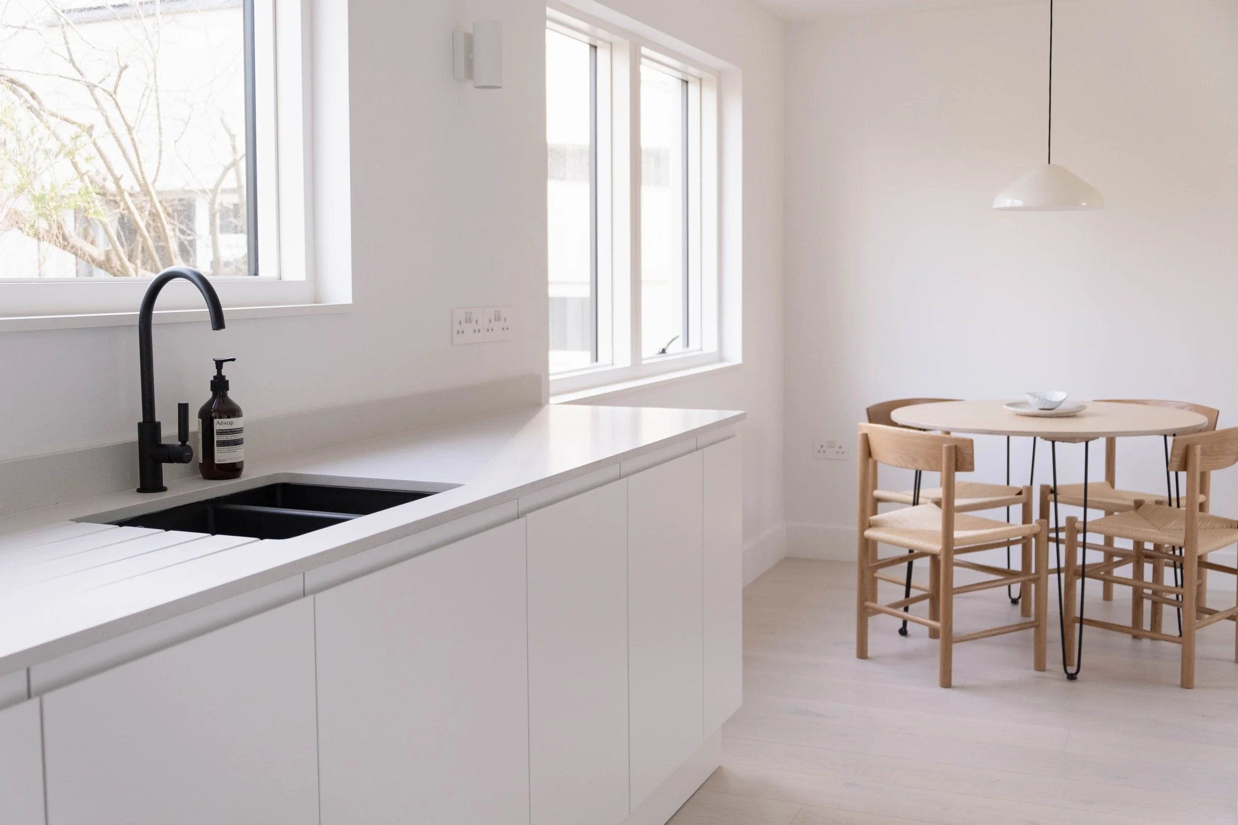 Bright white kitchen with black faucet, white countertop, and a bottle of hand soap. Adjacent dining area with light wooden table and matching chairs.