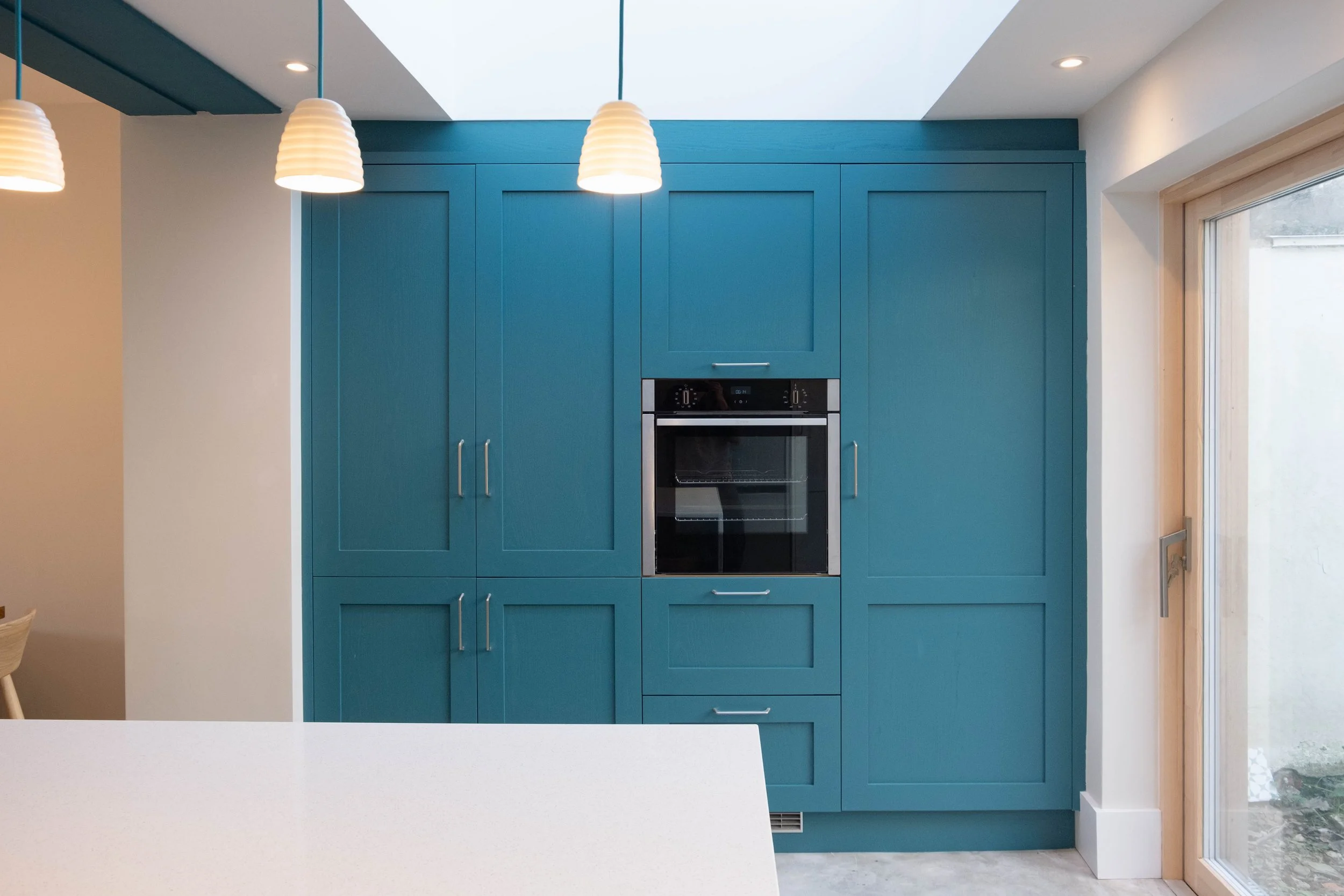 Modern kitchen with bright blue cabinetry, built-in oven, white countertop, ceiling lights, and a large window.