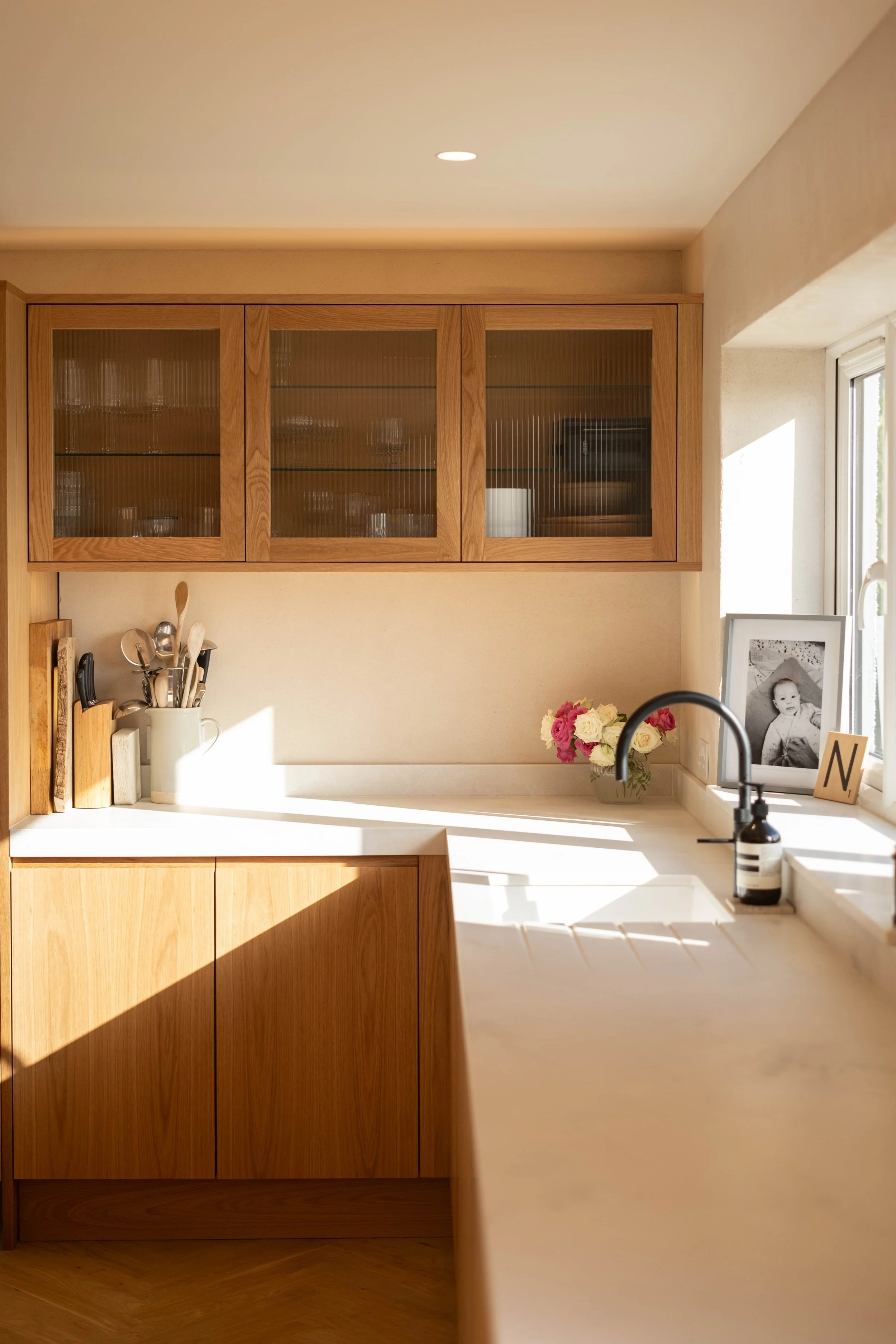 Kitchen with wooden upper cabinet with glass doors, white countertop, and black faucet, decorated with flowers and framed photos, near a window with sunlight.