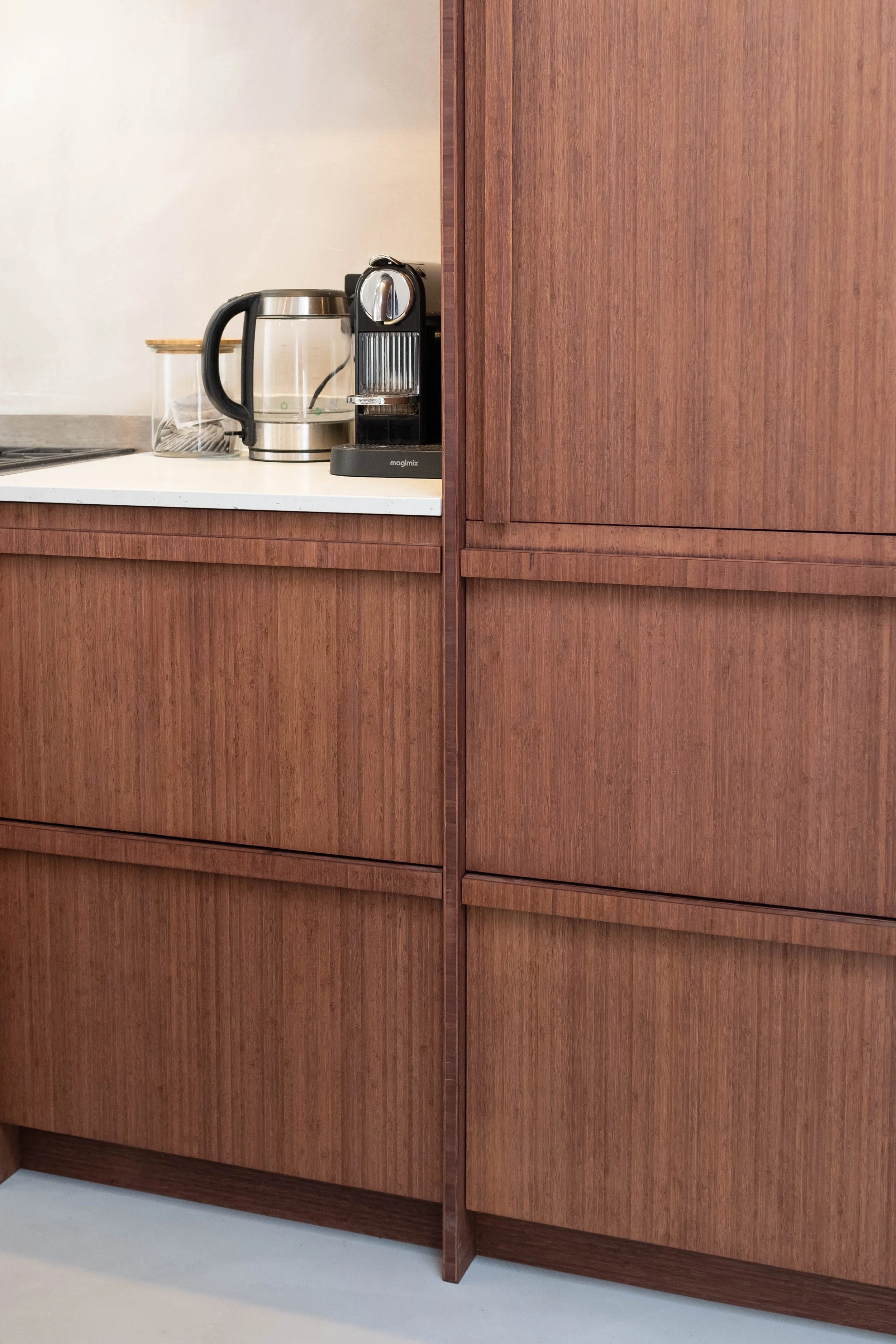 Interior view of a kitchen with wooden cabinets, a white countertop, a coffee machine, a glass jar, and a water kettle.