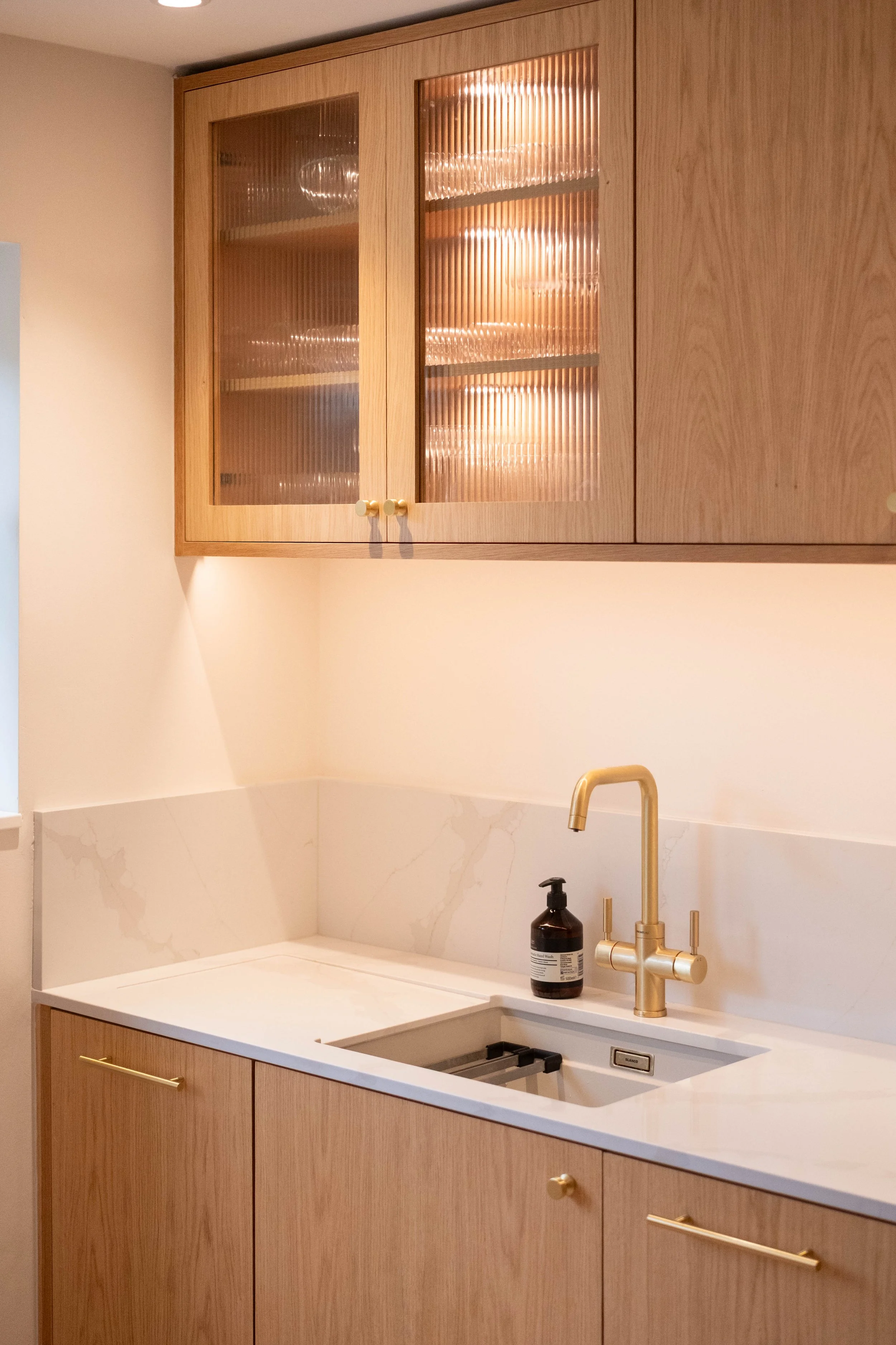 Kitchen with wooden cabinets, cream marble backsplash, and a white countertop. There is a built-in sink with a gold faucet and a black soap dispenser. The upper cabinets have glass doors with a ribbed texture, and gold knobs. There is a small section