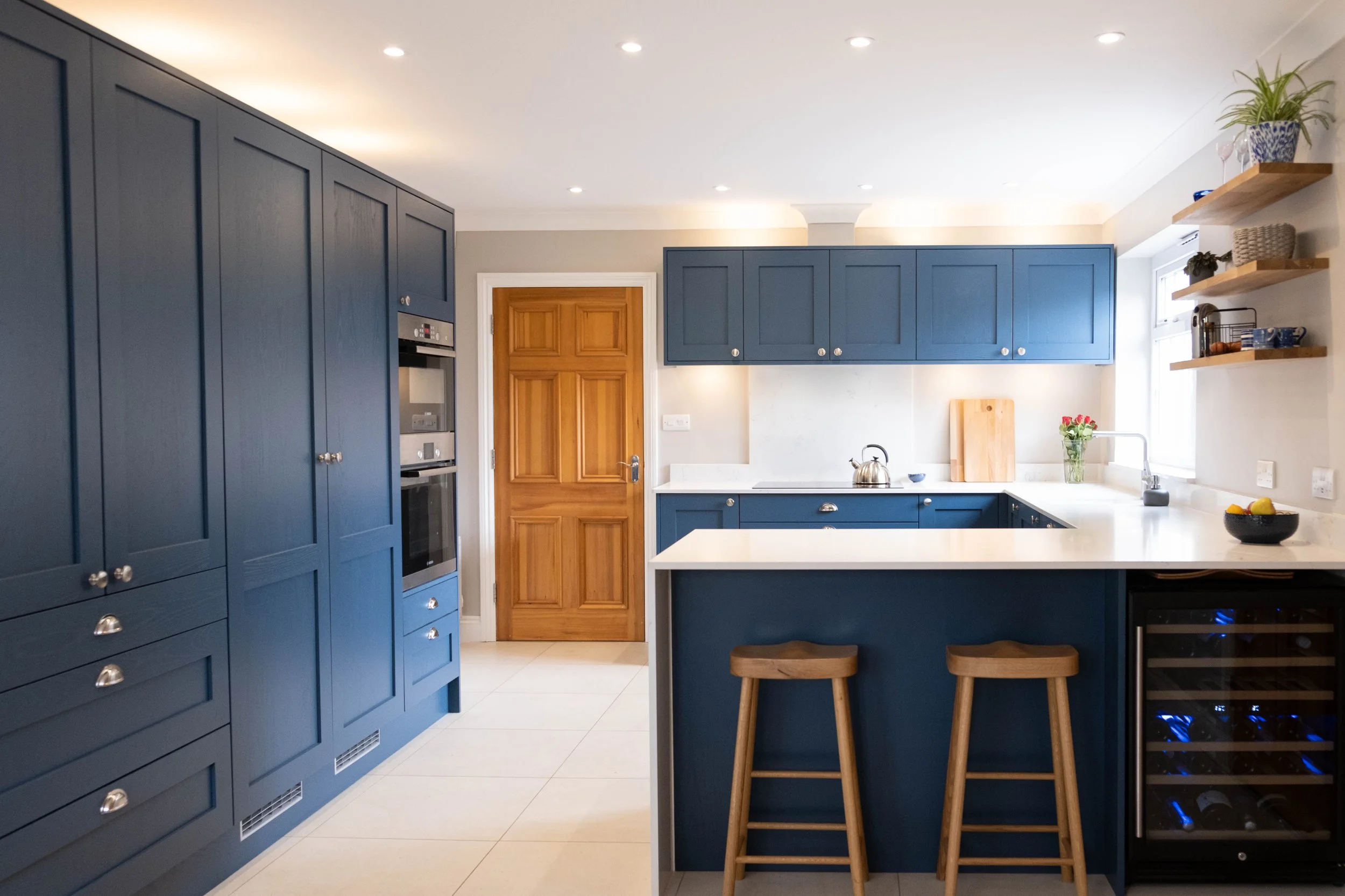 A modern kitchen with blue cabinets, a wooden door, white countertops, open wooden shelves with potted plants and bowls, and a small breakfast bar with two wooden stools.