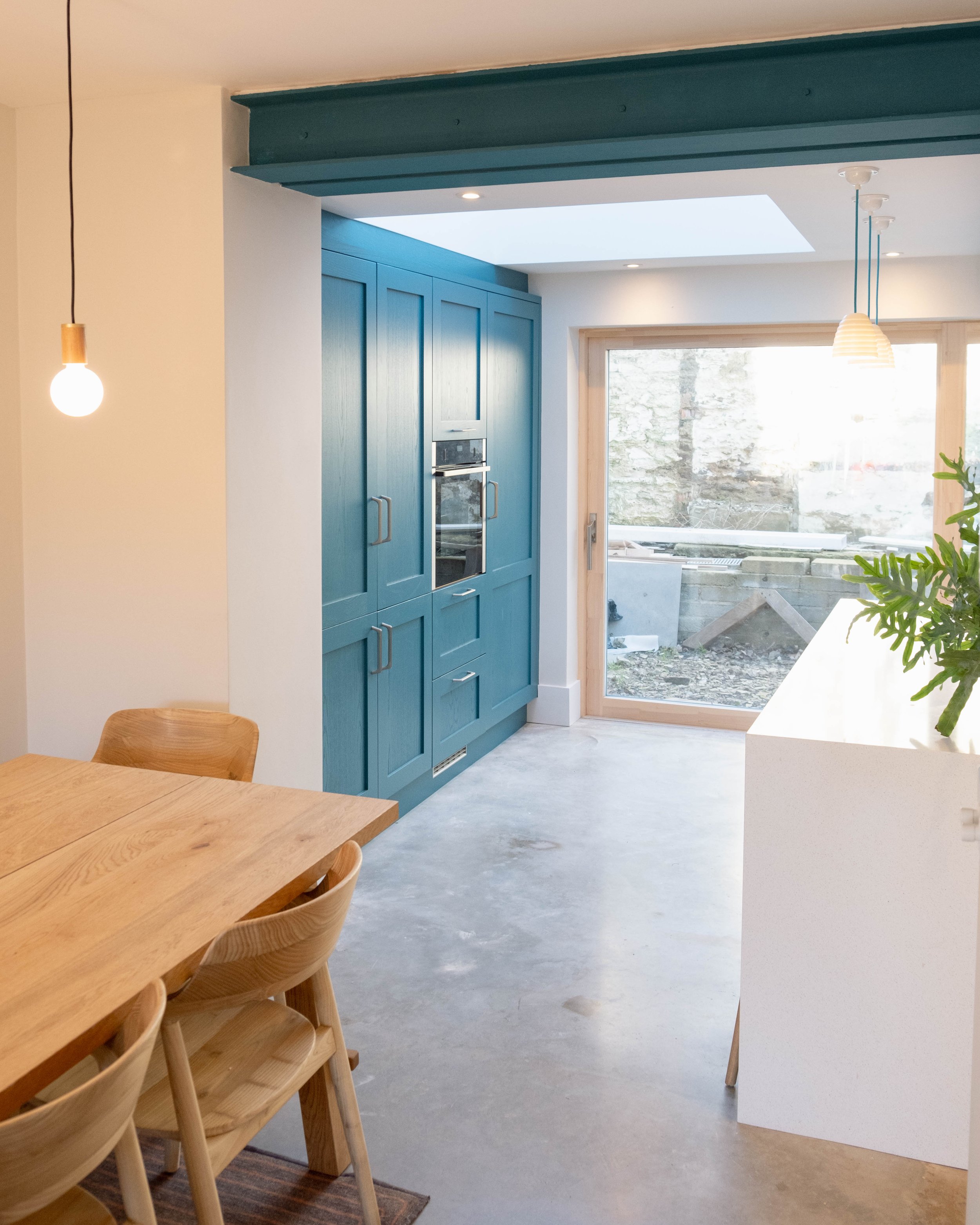 A modern kitchen with blue cabinetry, a large window, pendant lights, and a wooden dining table with matching chairs.