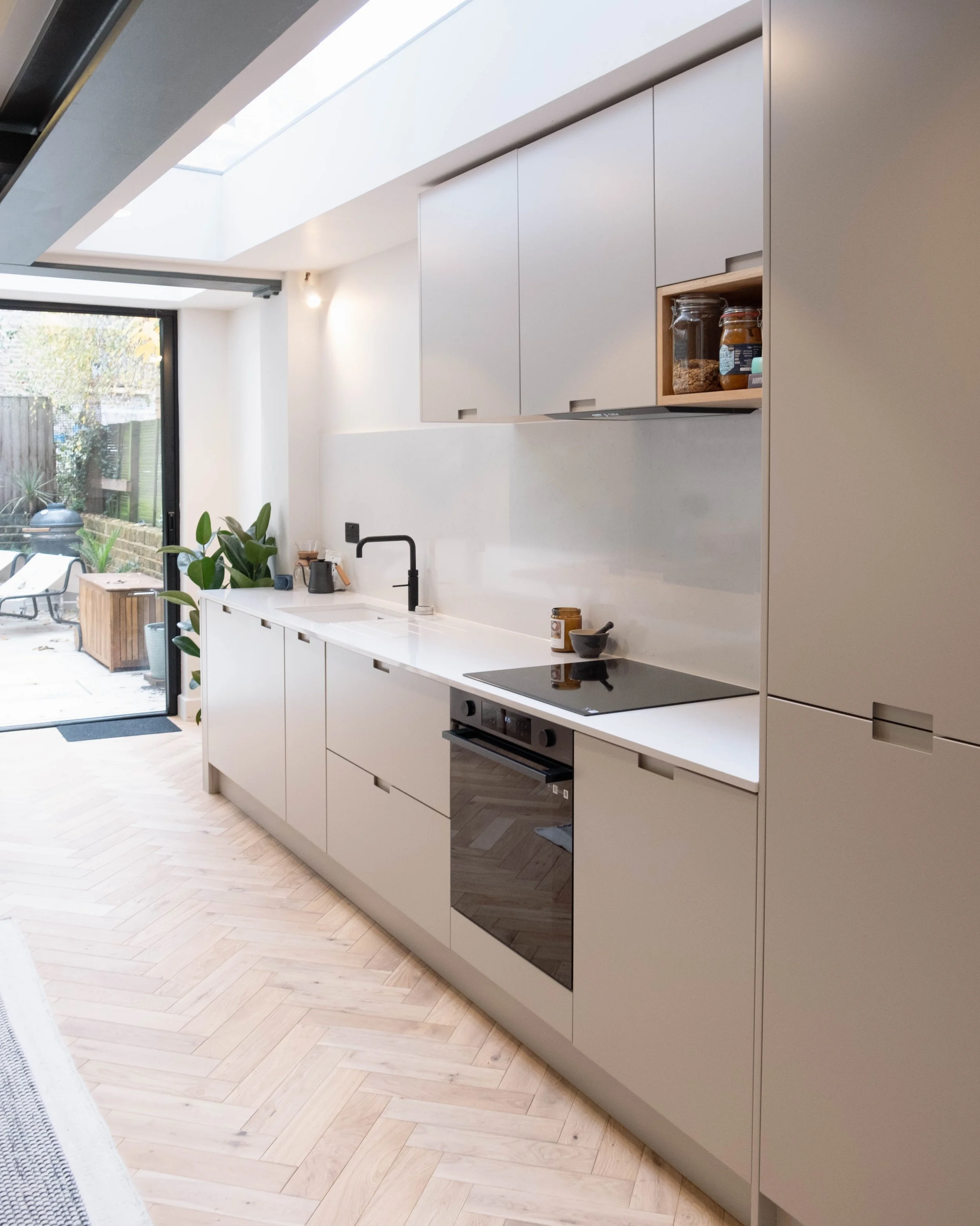 Modern kitchen with white cabinets, black stovetop, black faucet, light wood herringbone flooring, open to a backyard with outdoor seating and greenery, large ceiling skylight.