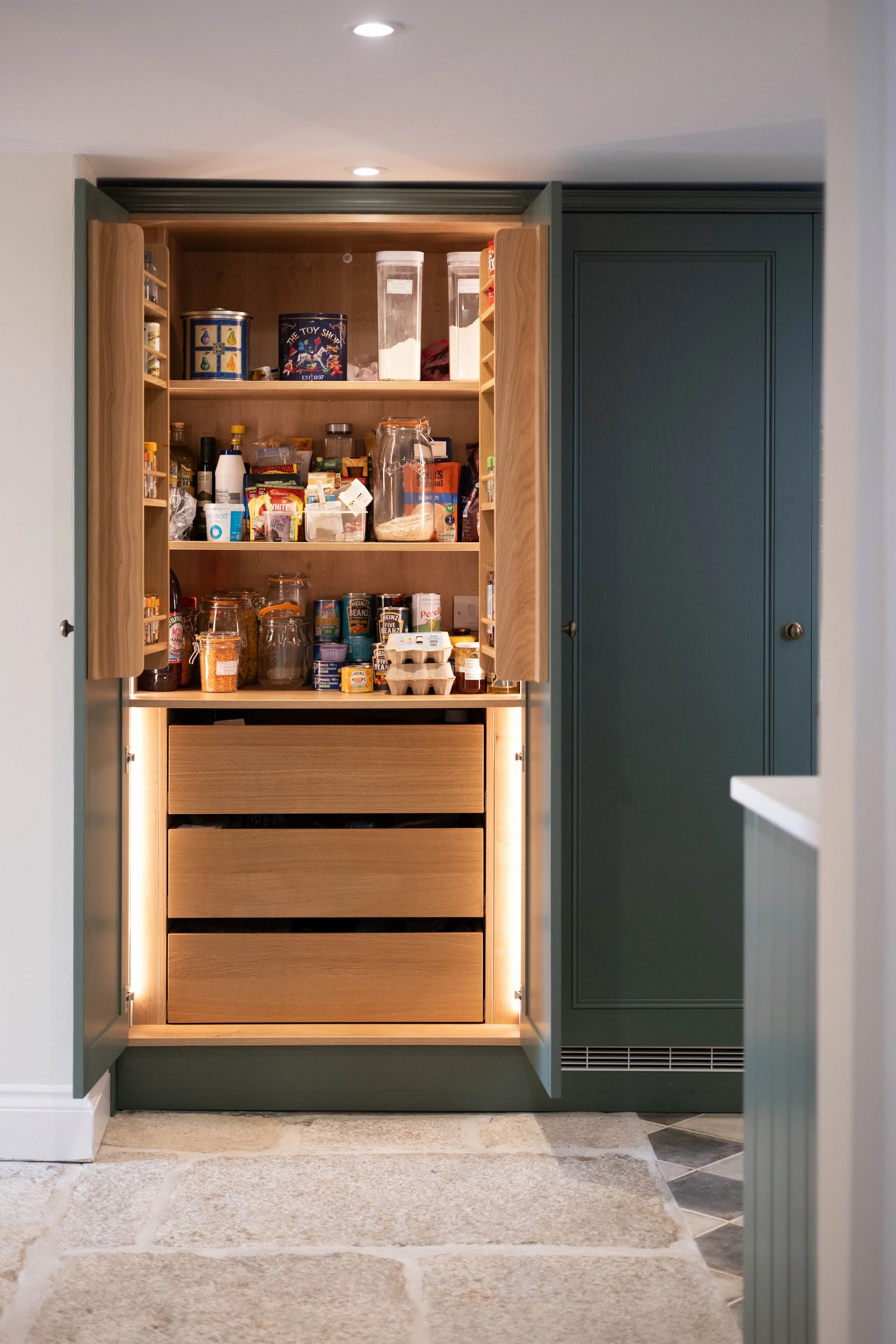 Open kitchen cabinet with shelves filled with jars, food containers, and pantry items, illuminated with built-in lighting, beside a teal-colored cabinet and a stone tiled floor.