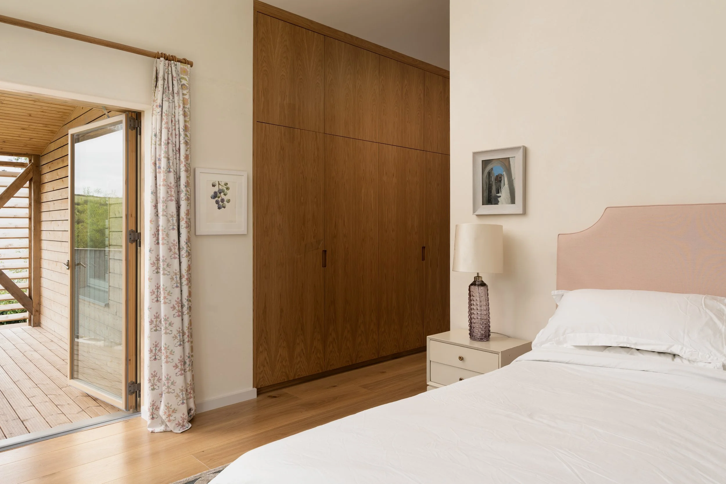A bedroom with a white bed, a nightstand with a purple lamp, a wooden wardrobe, framed wall art, and an open door leading to a wooden deck outside.