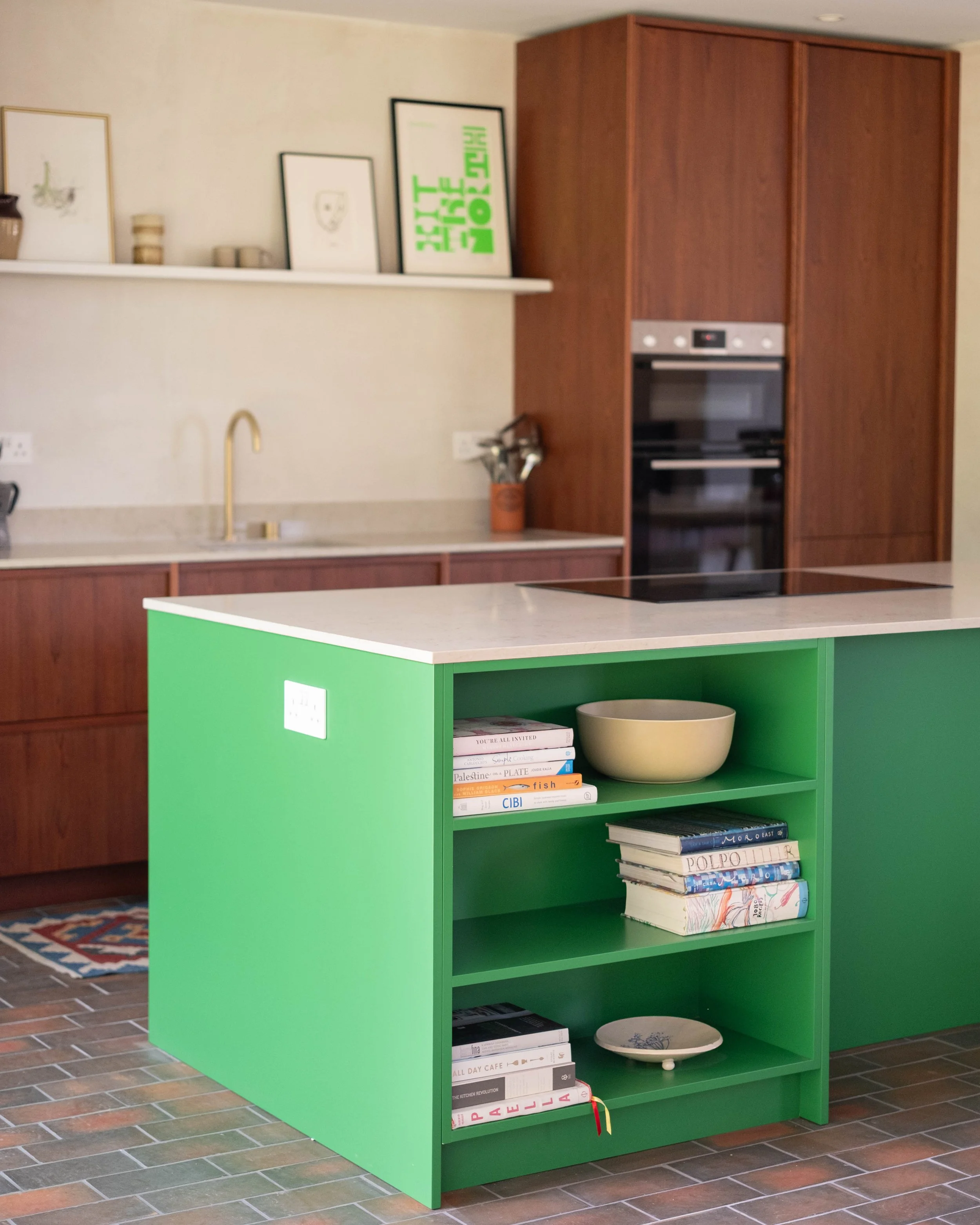 A kitchen with a green bookshelf filled with books and a decorative plate on the bottom shelf. The countertop has a beige bowl. In the background, there is a wooden cabinet with an oven, a white shelf with framed artwork and small decorative items, a