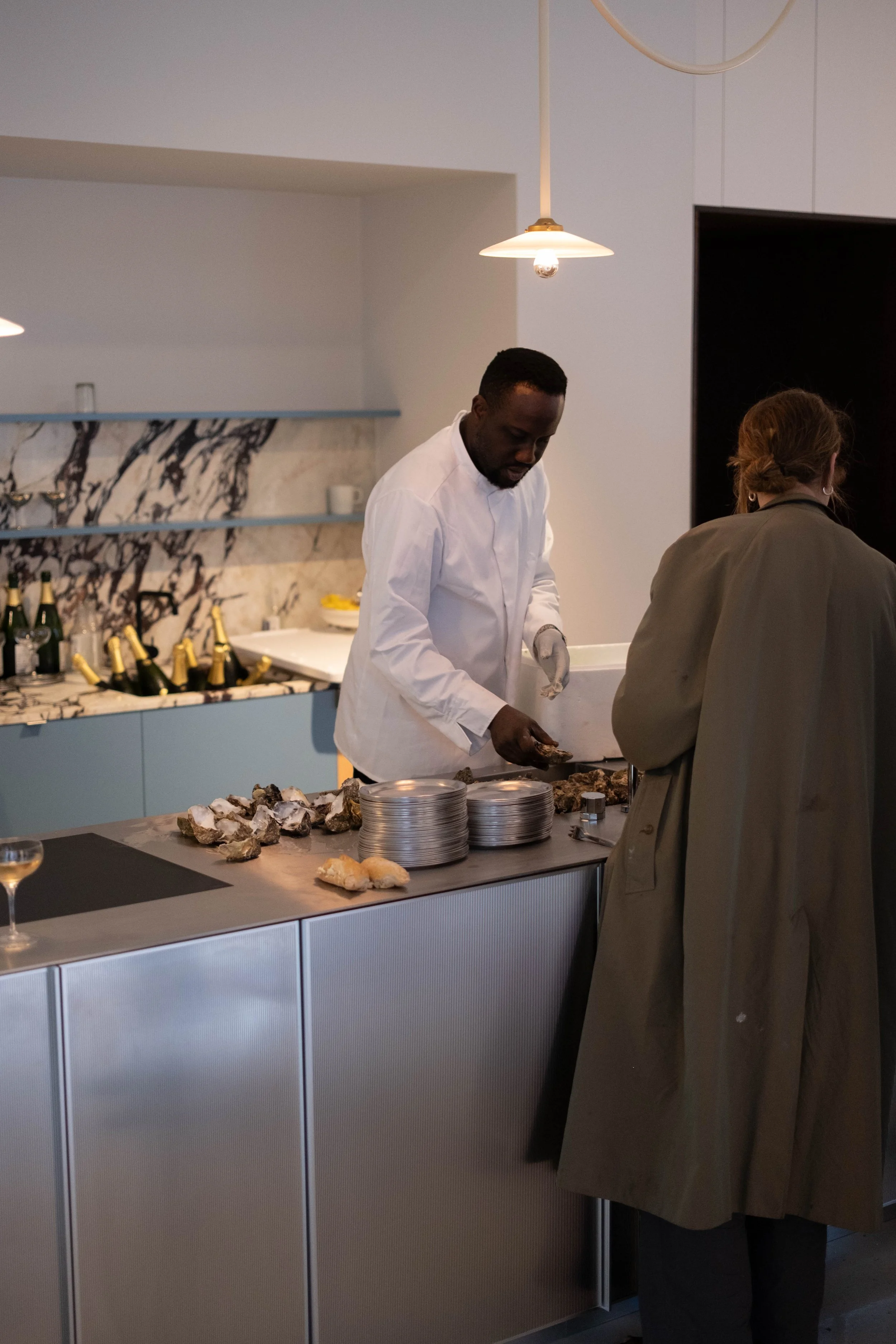 A man in a white chef's coat prepares food at a kitchen counter, with a woman in a gray coat standing nearby. The kitchen features a marble backsplash, open shelves with glasses, and bottles of wine or champagne.