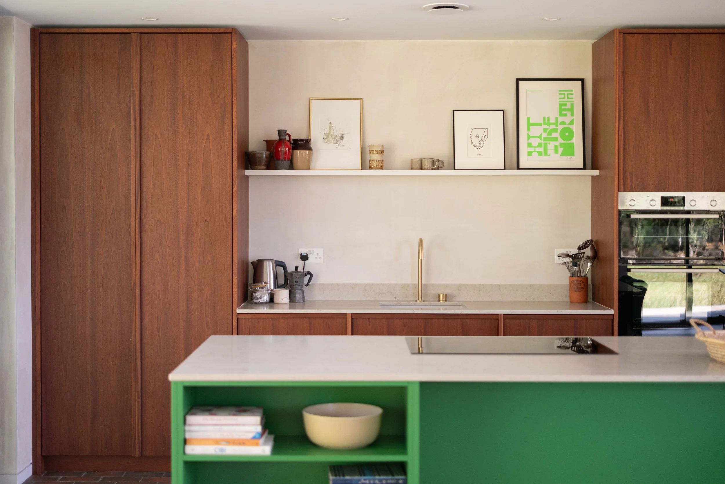 Modern kitchen with wooden cabinets, a white countertop, and a green island with books and a yellow bowl. Decor includes framed artwork, vases, mugs, and utensils.