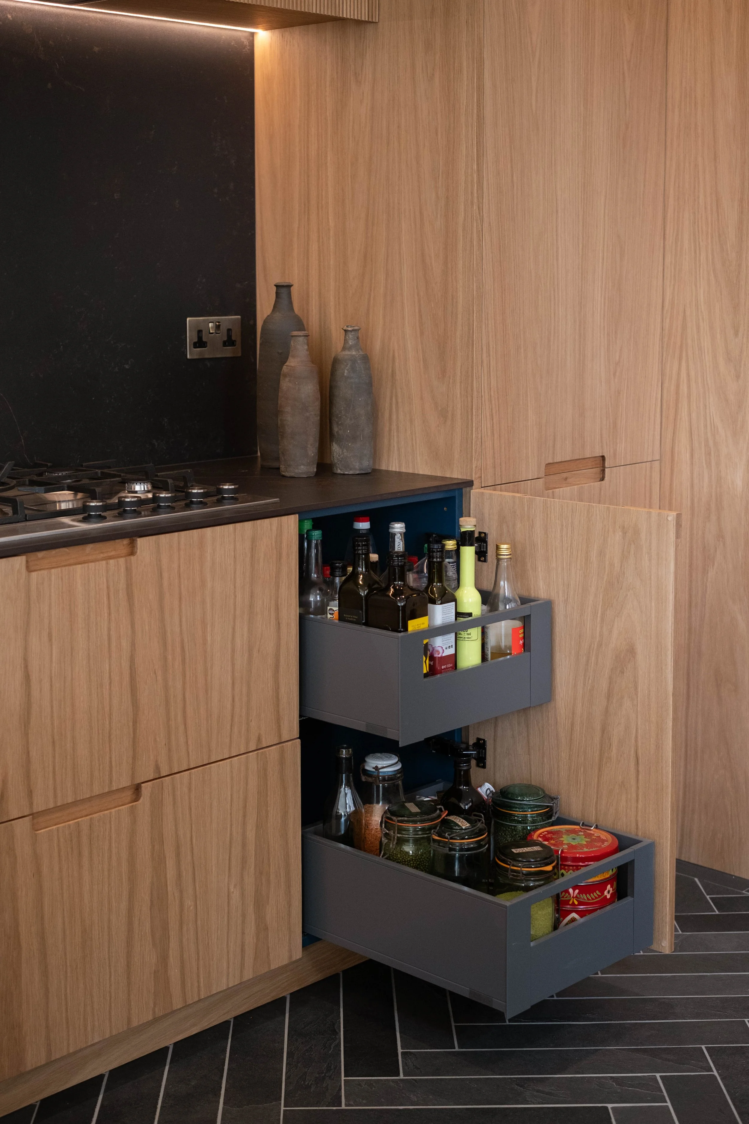Open drawer in a kitchen with wooden cabinets, containing various bottles, jars, and tins, with a black backsplash and a black and gray tiled floor.