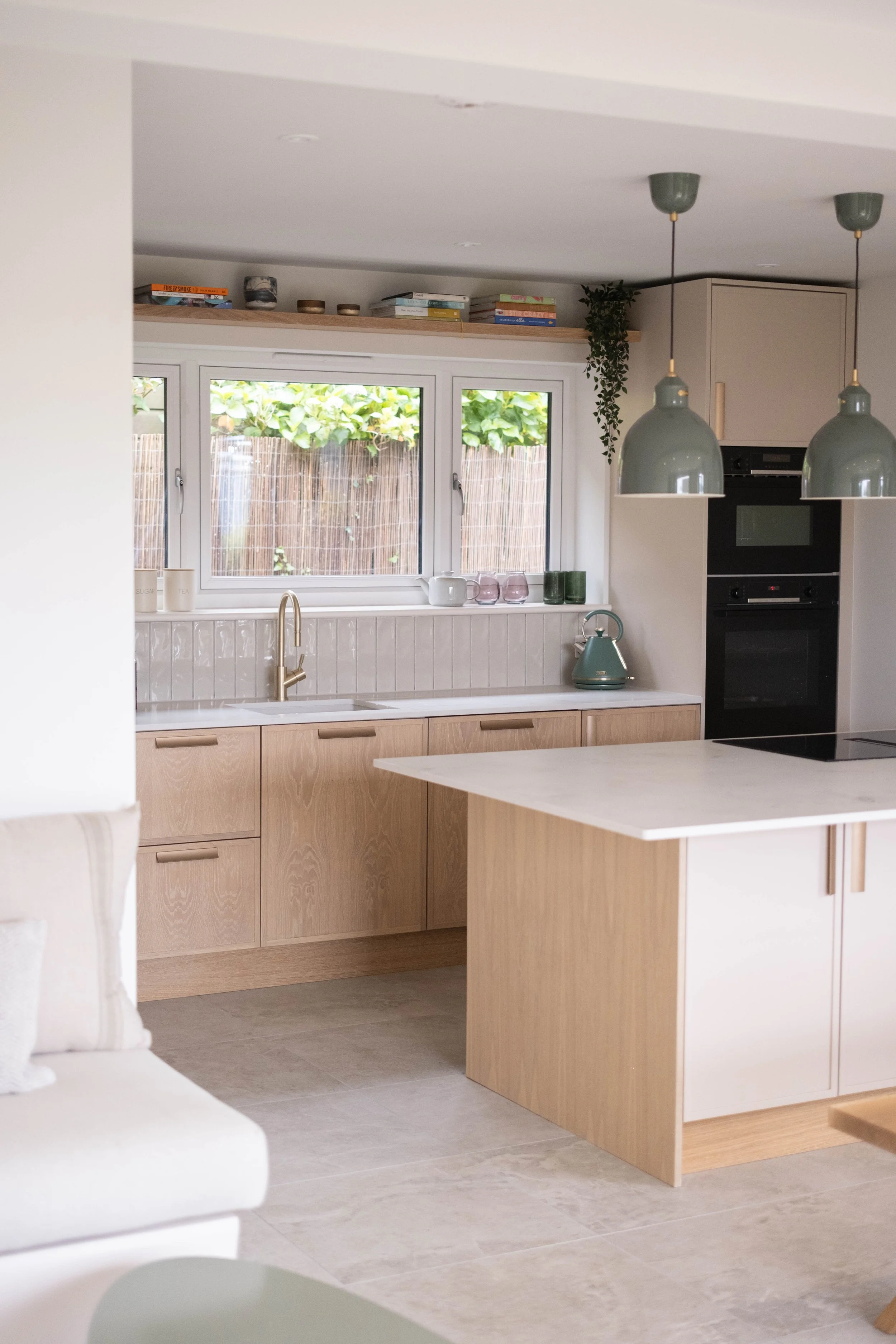 Modern kitchen with beige cabinets, a white countertop, and a window with greenery outside. Two pendant lights hang from the ceiling, and a small countertop with a teal kettle is visible.