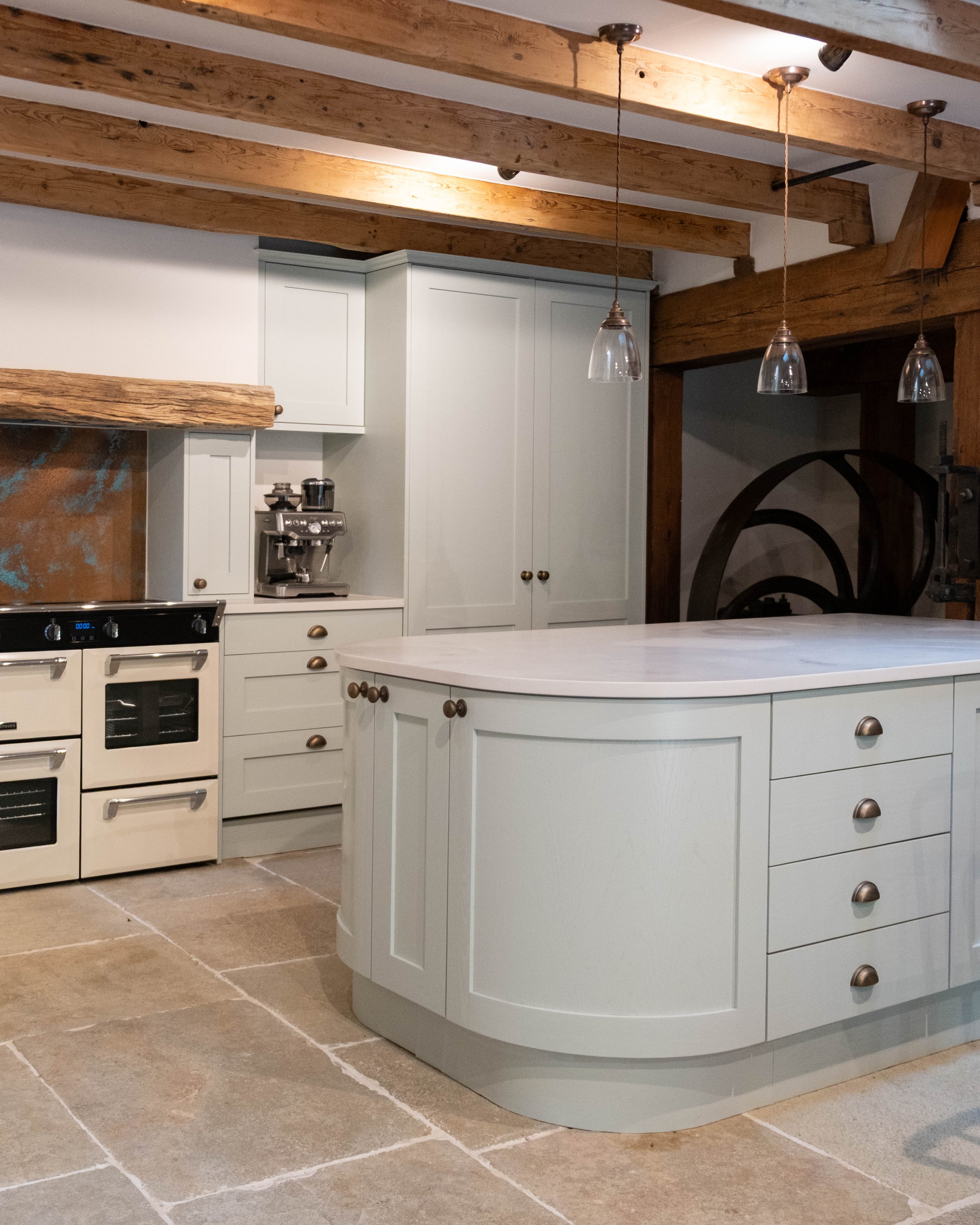 A kitchen with rustic wooden beams on the ceiling and a white kitchen island with drawers. The flooring is tiled with large, brownish tiles. There are hanging pendant lights above the island, and a coffee machine is visible on the counter.