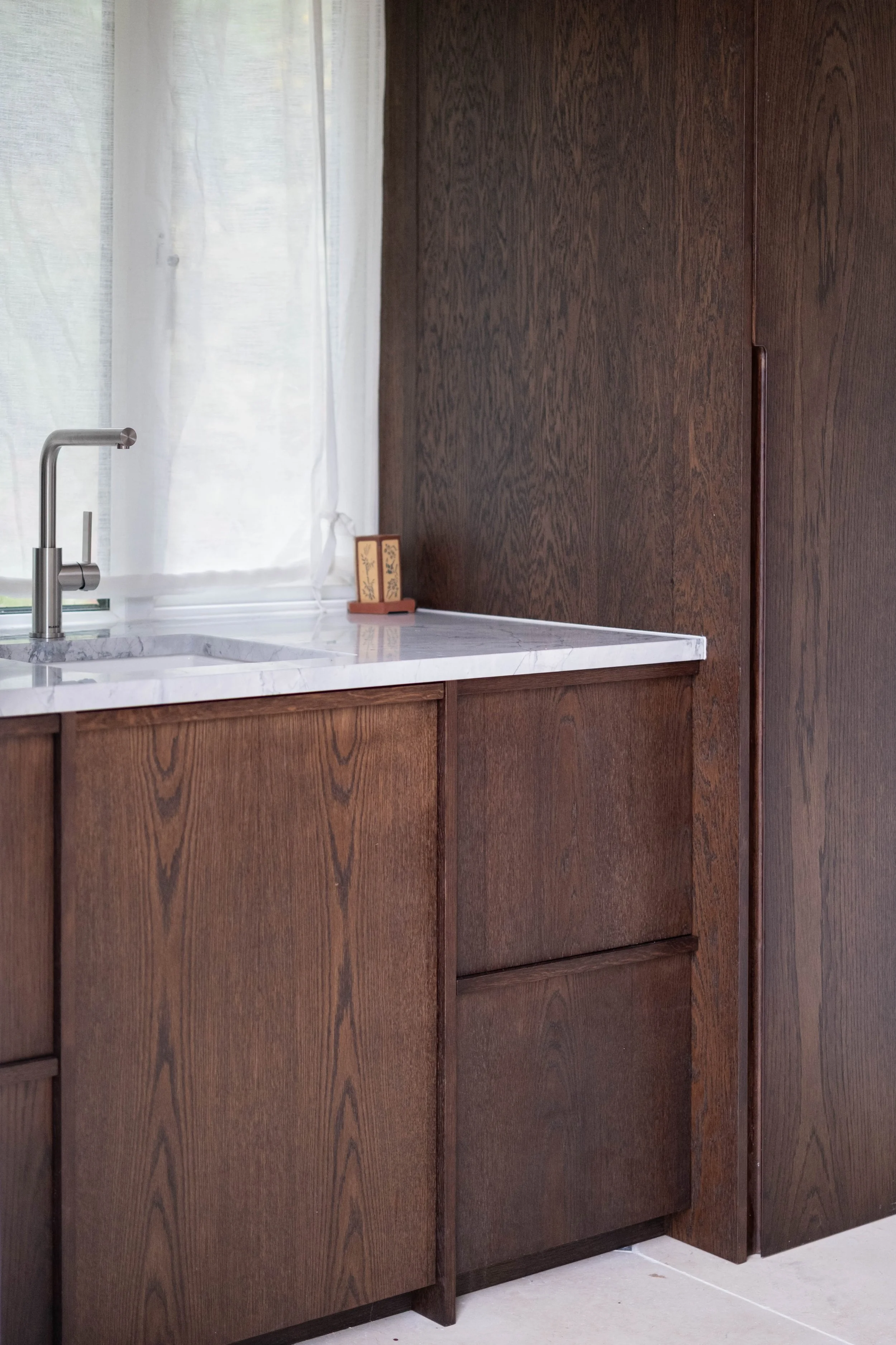 Kitchen with dark wooden cabinets, white marble countertop, silver faucet, white curtains, and a small decorative item on the counter.