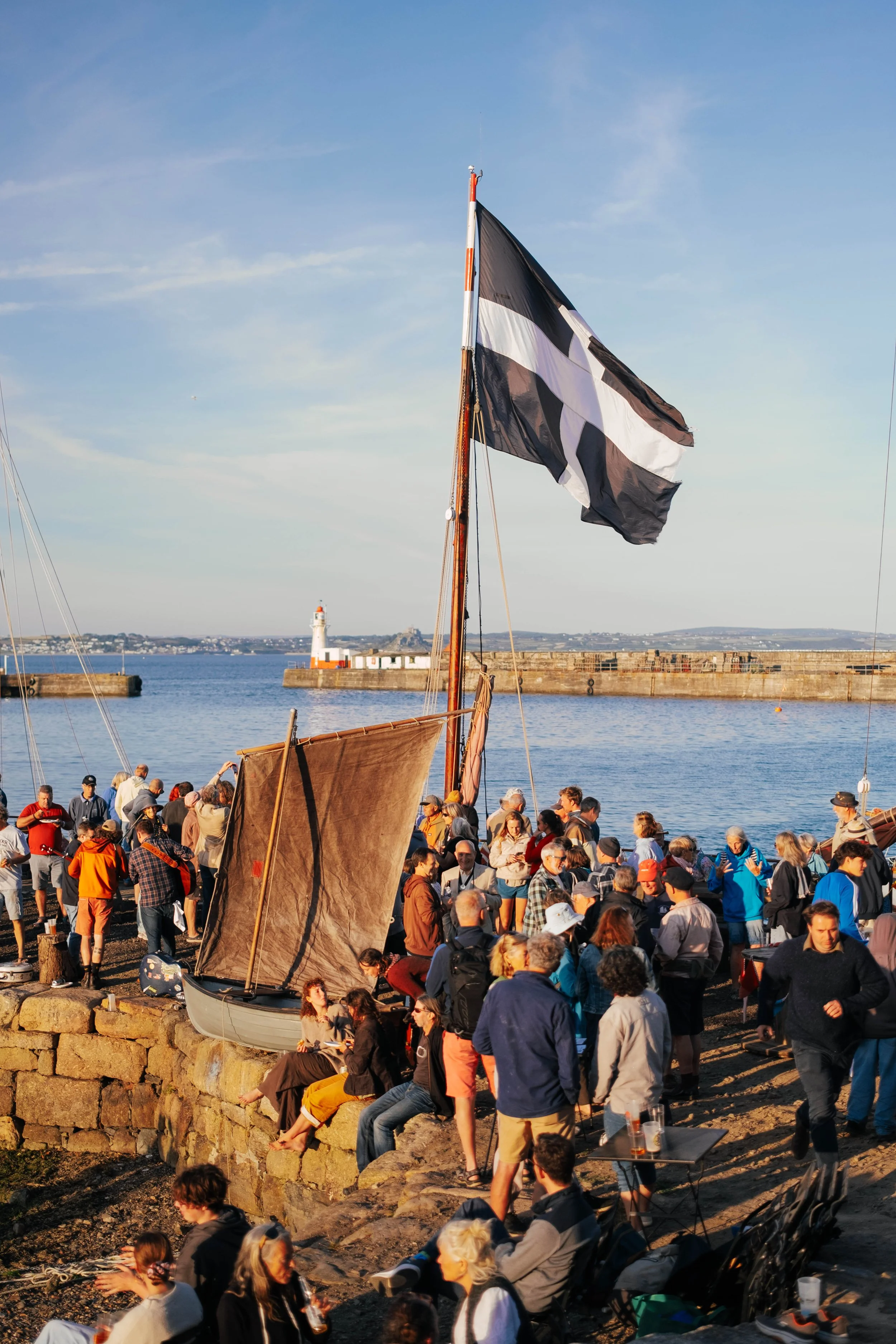 A large crowd of people gathered near a marina with boats and a lighthouse in the background, during the daytime.