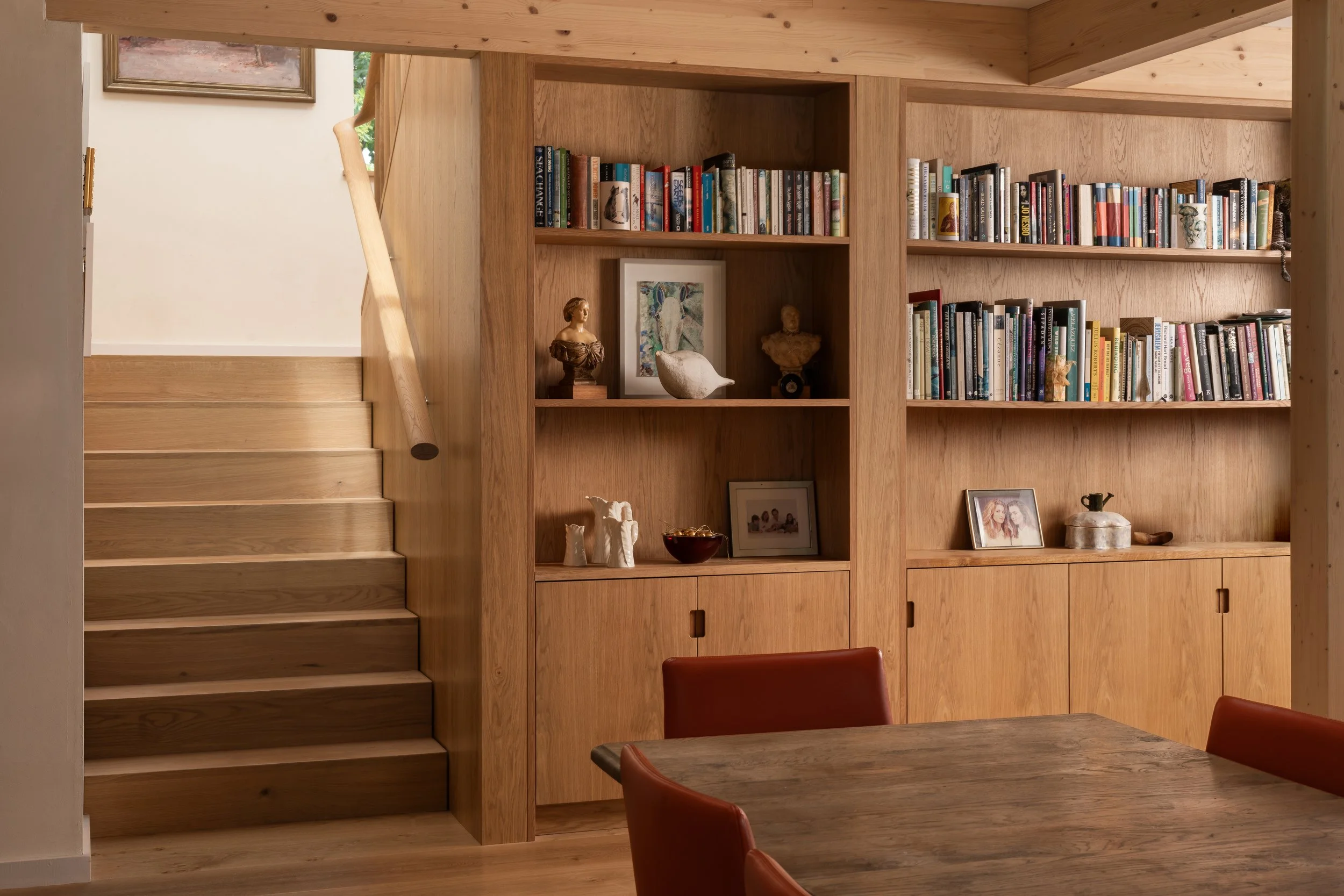 Wooden bookshelf against wood-paneled wall with books, framed pictures, and decorative items, next to a staircase and table with leather chairs.