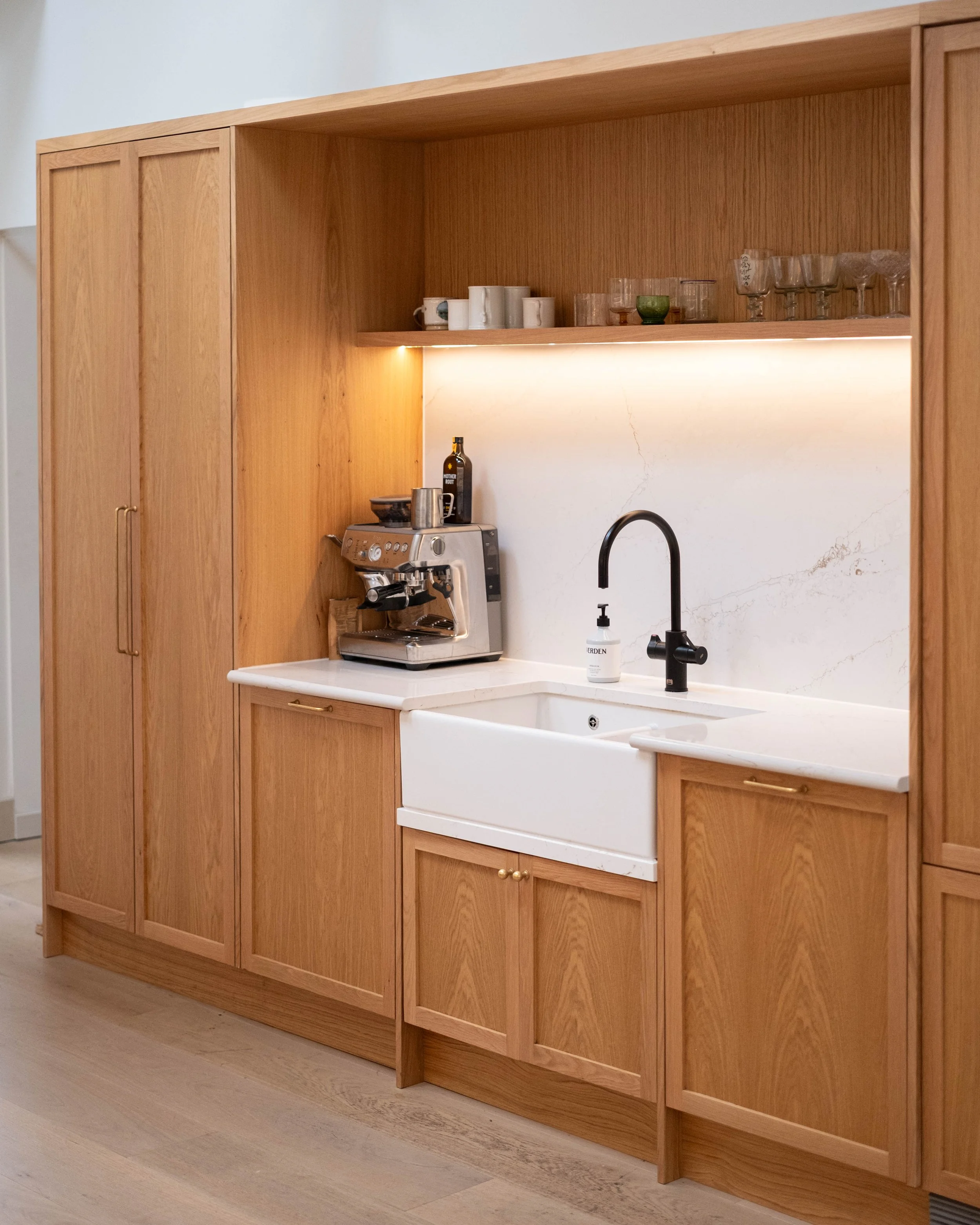 A wooden kitchen cabinet with a white marble countertop and a black kitchen faucet. An espresso machine is on the countertop, and shelves above hold various cups and glassware. The background features a white marble backsplash with subtle veining.
