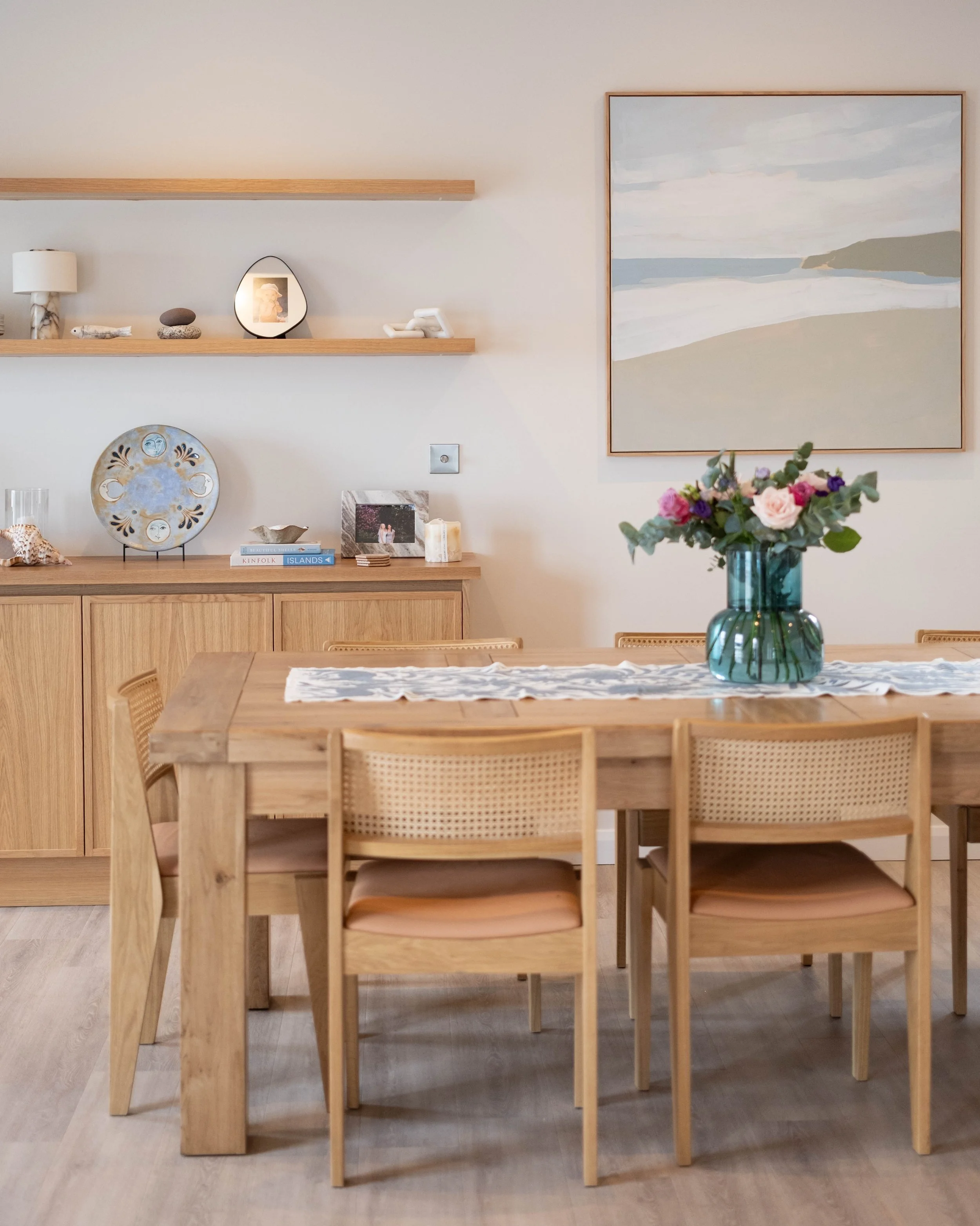 A dining room with a wooden table and six matching chairs with rattan seats. On the table is a green glass vase with pink, purple, and white flowers. The background includes a wall-mounted wooden shelf with decorative items, a large landscape paintin