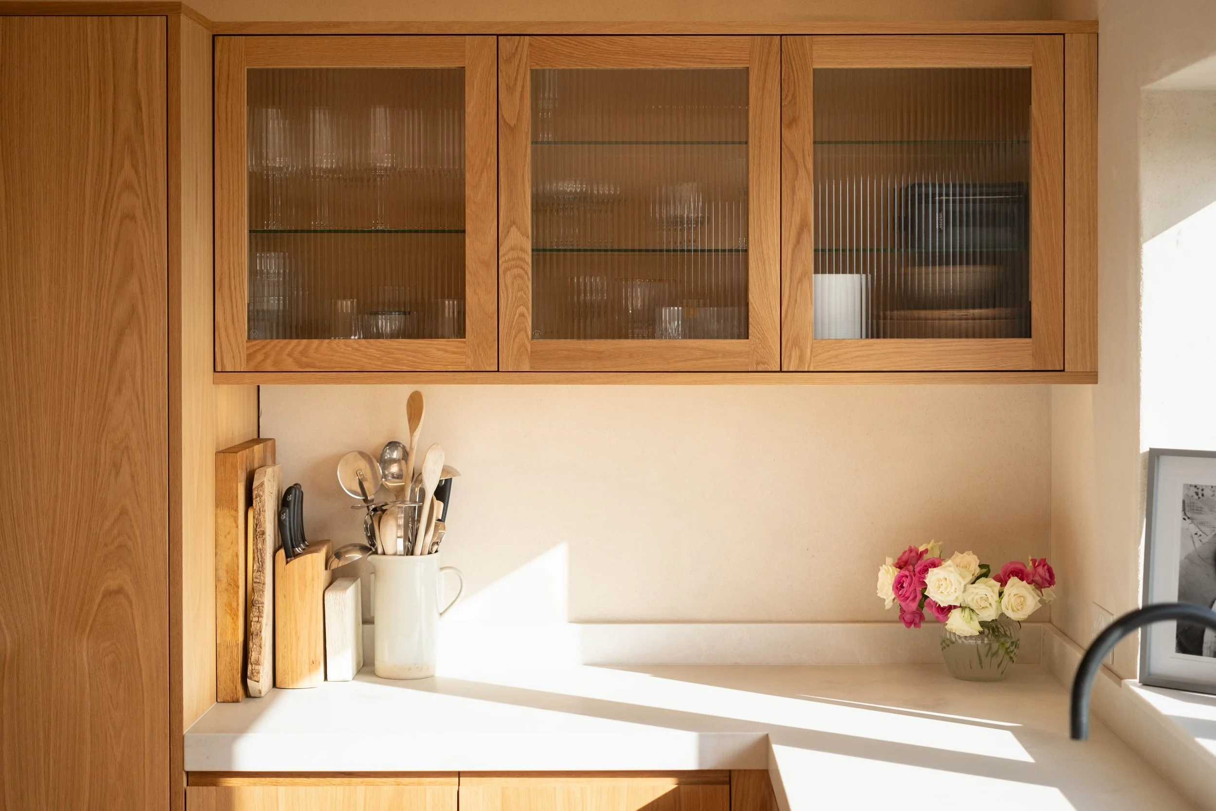 Wooden kitchen cabinet with glass doors, containing glassware inside. Below the cabinet, a countertop holds a white pitcher, and a collection of kitchen utensils in a white container. To the right, a vase with pink and white flowers is visible, and a