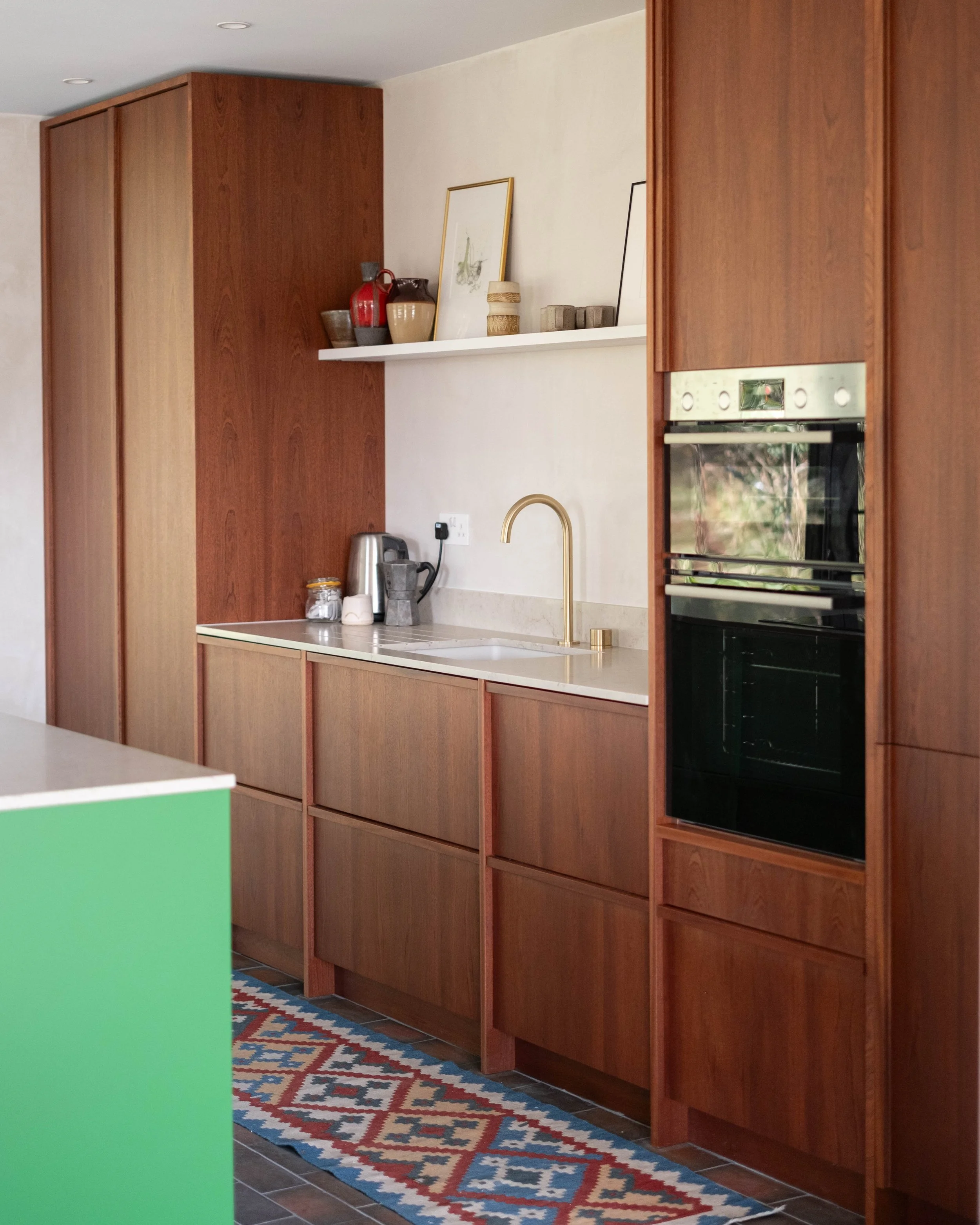 A kitchen with wooden cabinets, a white countertop, and a colorful geometric patterned rug on the floor. There are decorated vases and framed pictures on a white shelf above the counter, and a brass kitchen faucet over the sink.