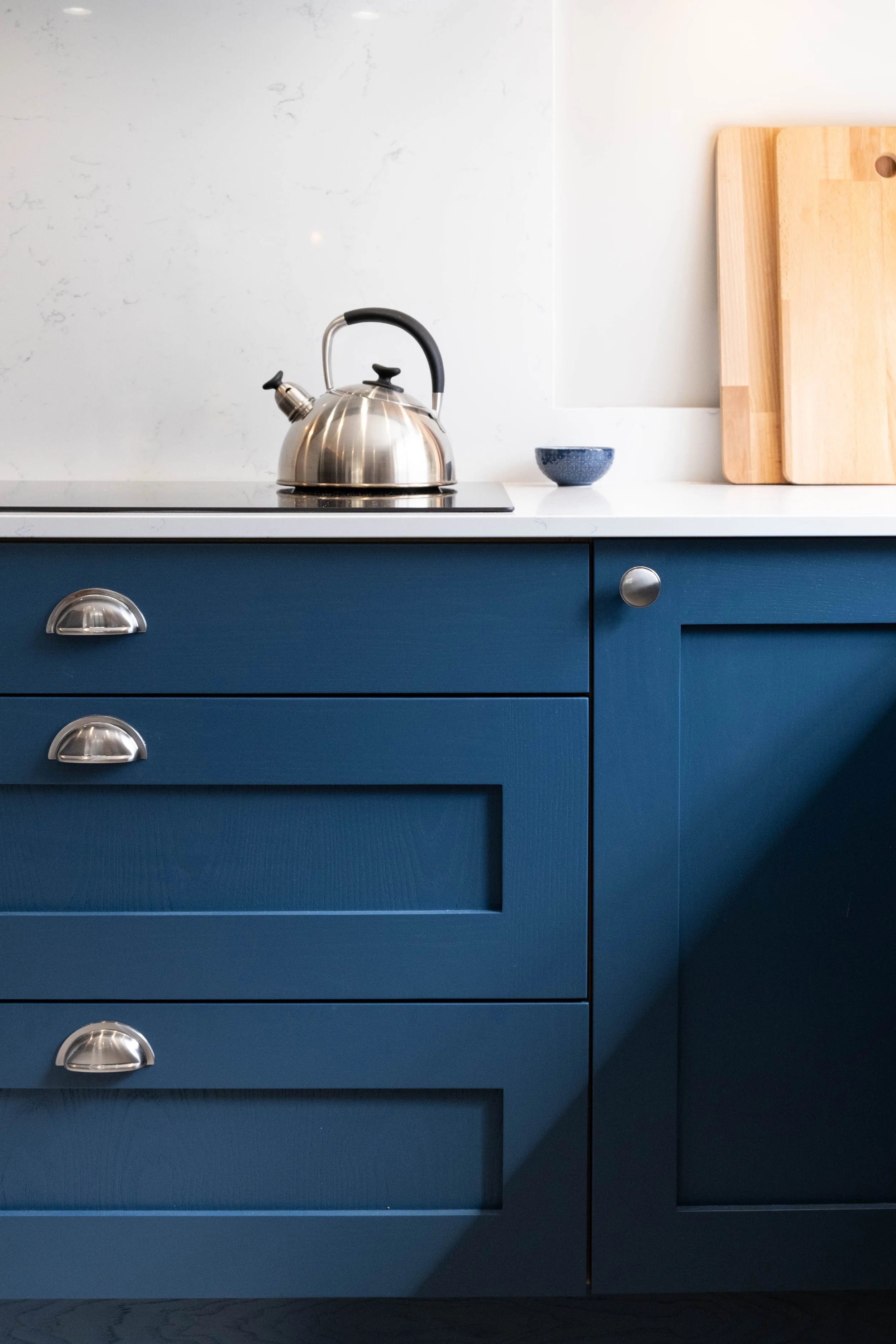 Modern kitchen with navy blue cabinetry, a stainless steel kettle on the stovetop, a small blue bowl, and two wooden cutting boards against a white wall.
