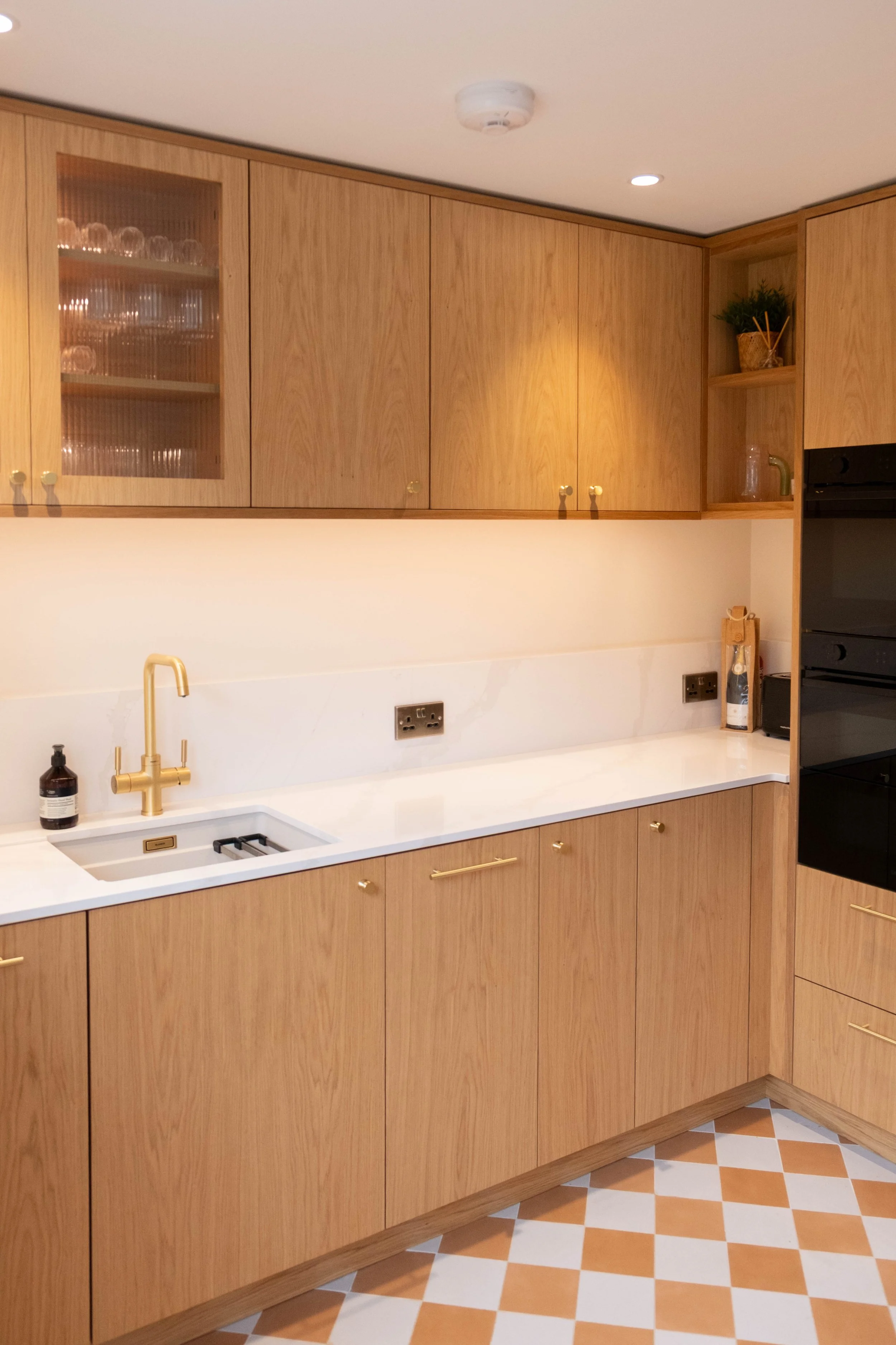 Kitchen with wooden cabinets and white countertop, gold fixtures, patterned floor tiles, including a sink with a gold faucet, bottles, and decor on the counter, and glasses stored in a glass-front cabinet.