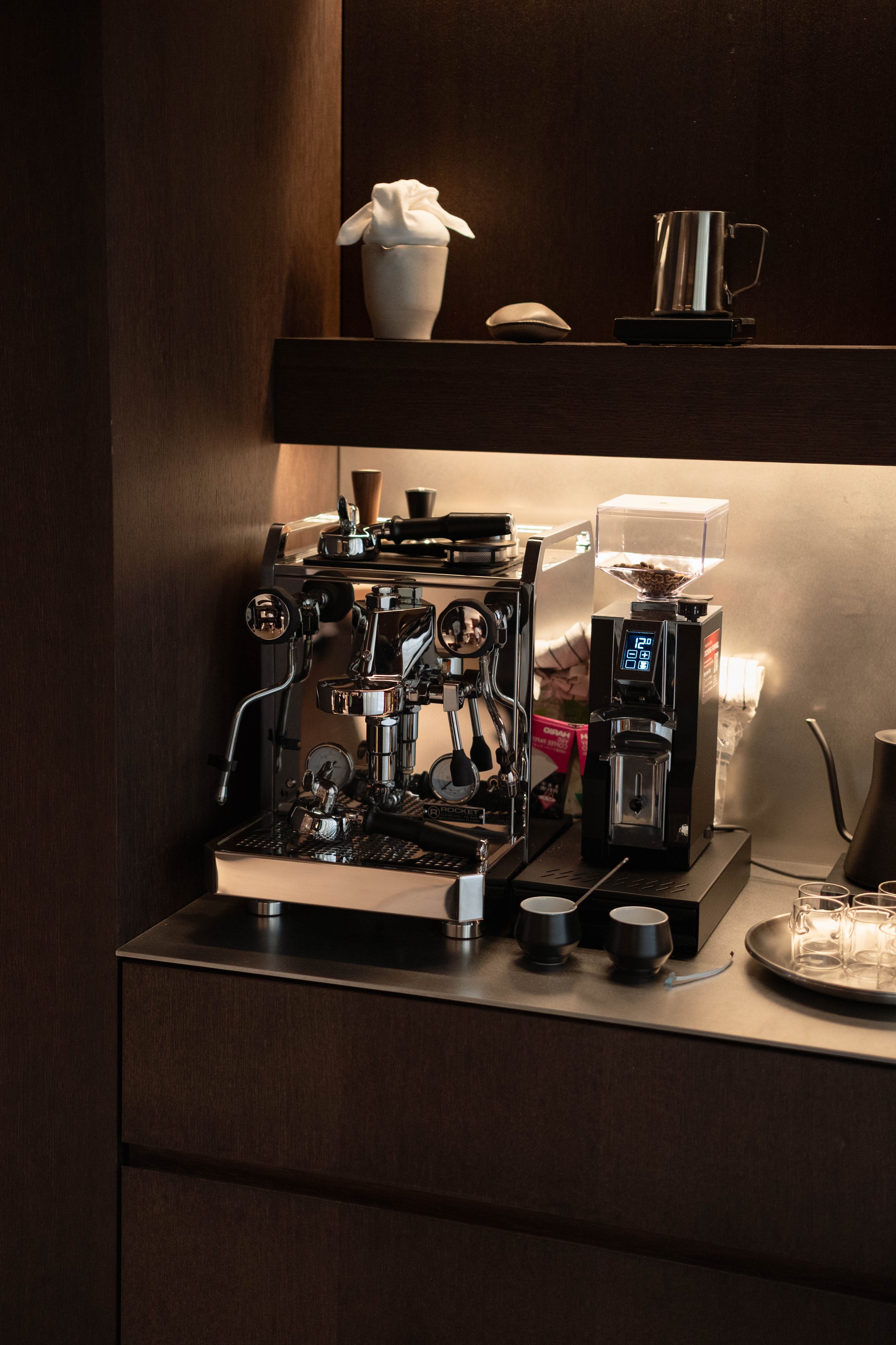 A coffee station with a professional espresso machine, coffee grinder, and various cups and utensils on a counter and shelf.
