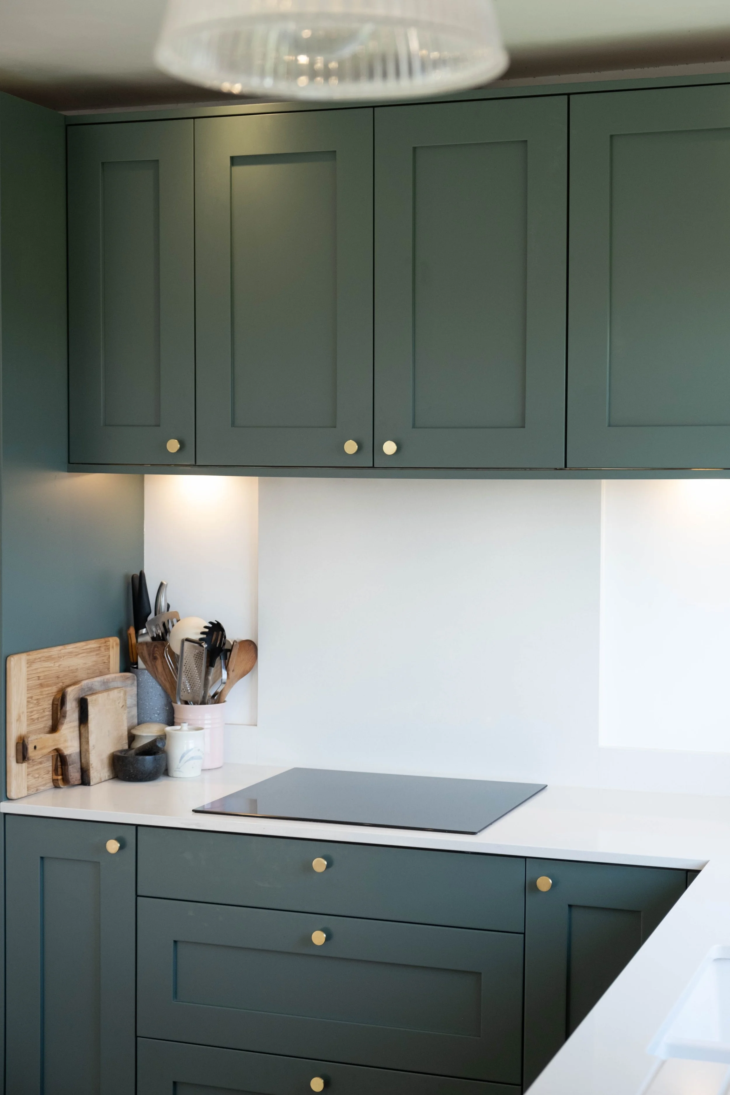 Green kitchen cabinets with round gold knobs, white countertop, and kitchen utensils including cutting boards, a strainer, and cups. Part of a stove is visible.