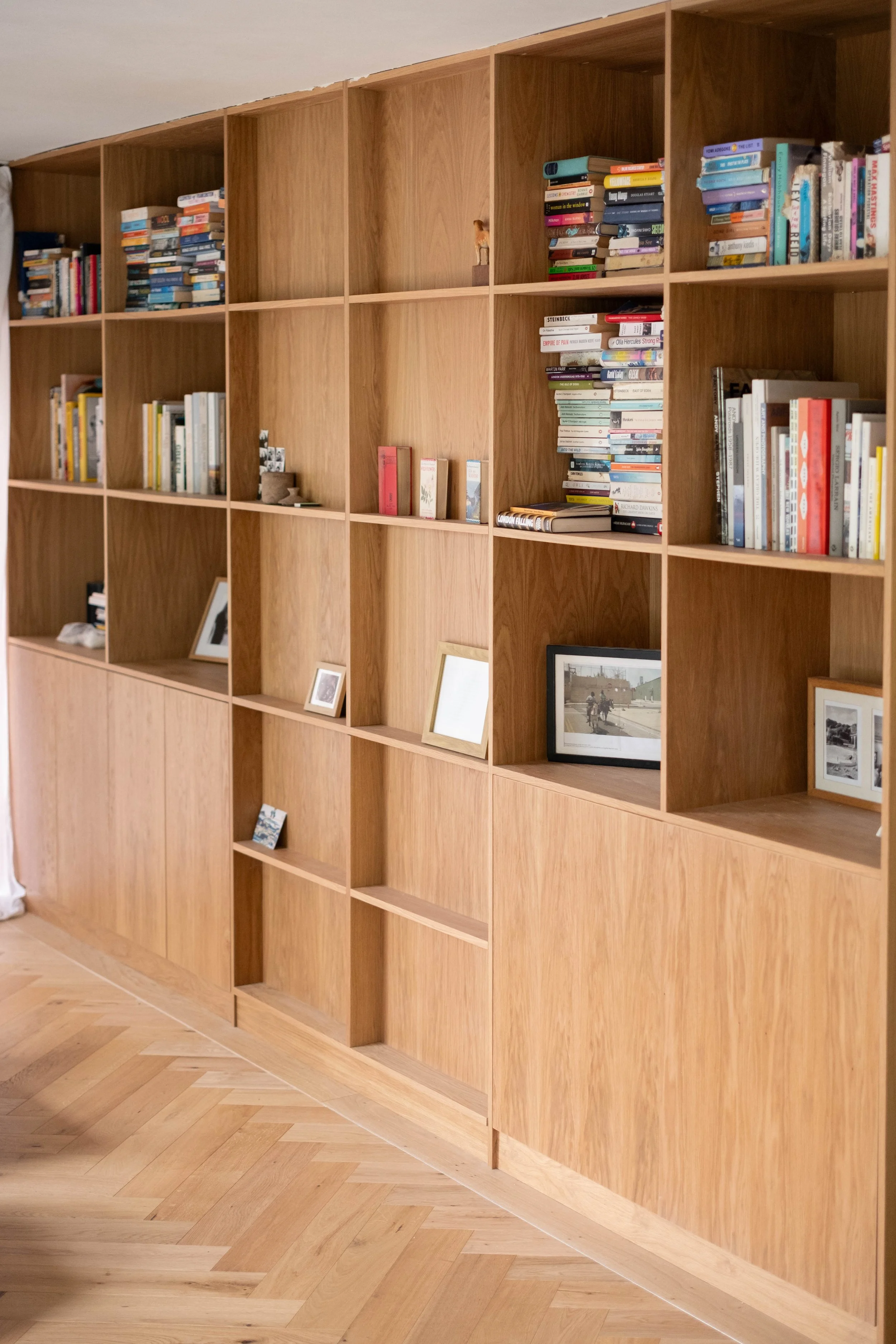 A large wooden bookshelf with multiple compartments, some containing books and framed photos, set against a light-colored wall and a herringbone wood floor.