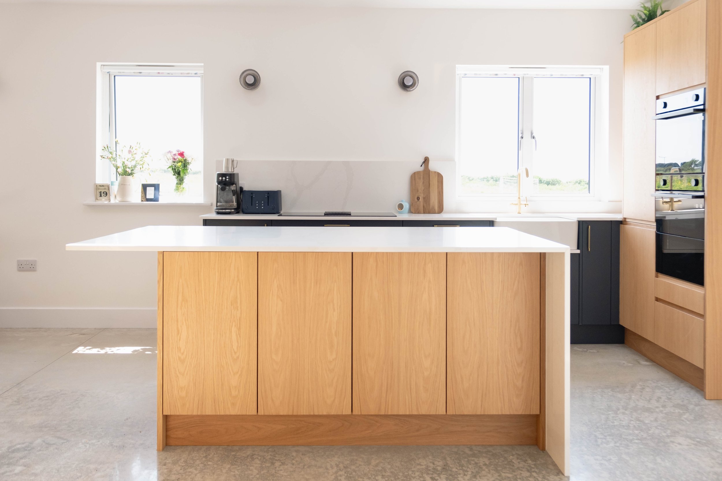 Modern kitchen with wooden cabinets, white countertops, two windows, plants, flowers, and kitchen appliances.